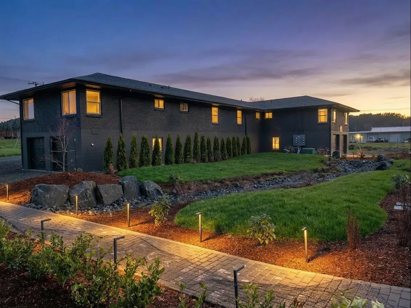 Commercial landscaping and management in downtown Florence, Oregon. Modern, two-story black house with lit windows, surrounded by a landscaped yard with a grassy lawn, small plants, large rocks, and pathway lights at dusk.