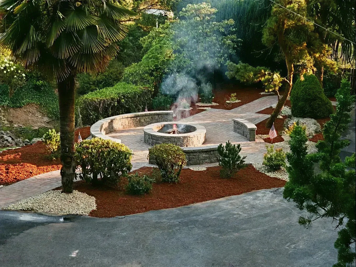 A landscaped backyard with a circular stone fire pit emitting smoke, surrounded by a paved patio, lush trees, bushes, and small American flags in Dunes City, Oregon