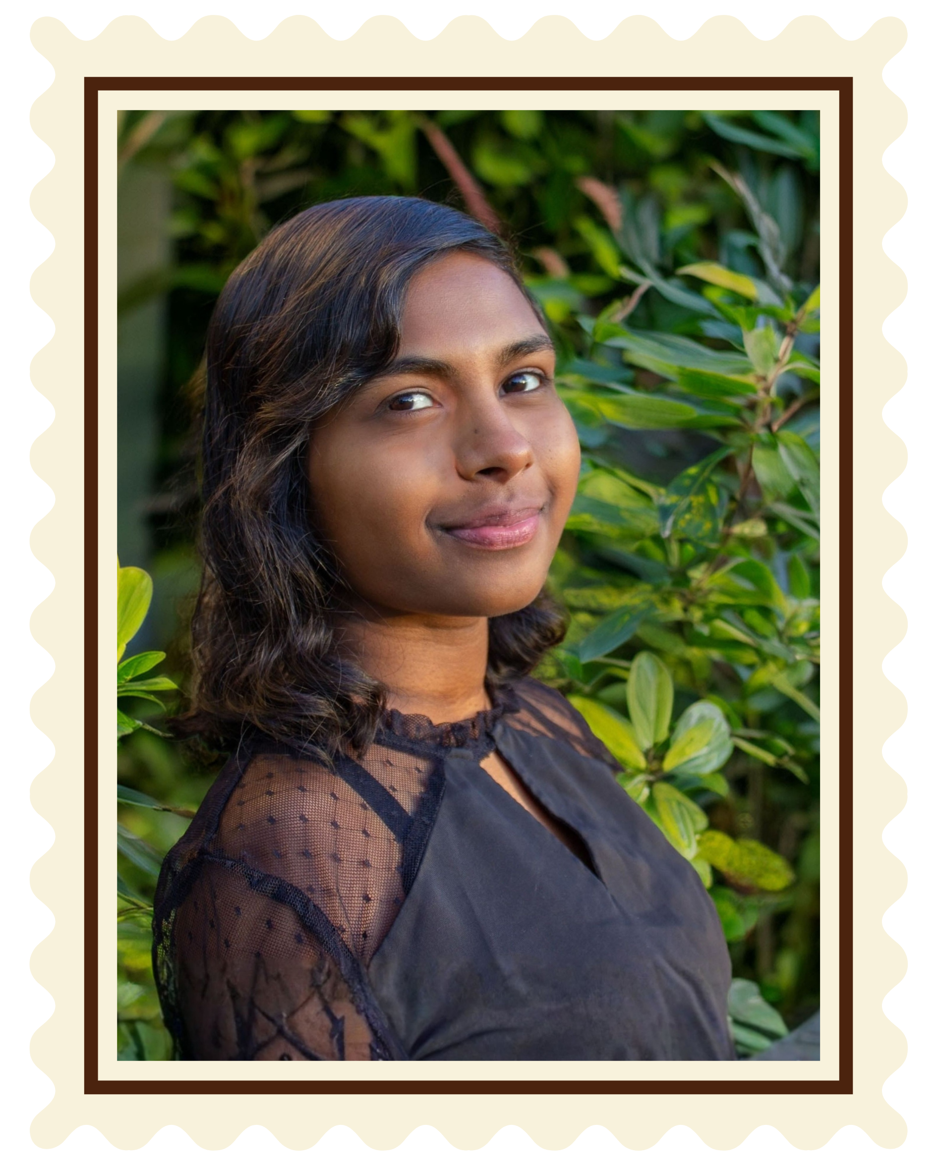 A portrait of a young woman with shoulder-length wavy hair, standing outdoors among green leafy plants, wearing a black blouse with sheer polka dot sleeves, smiling gently at the camera.