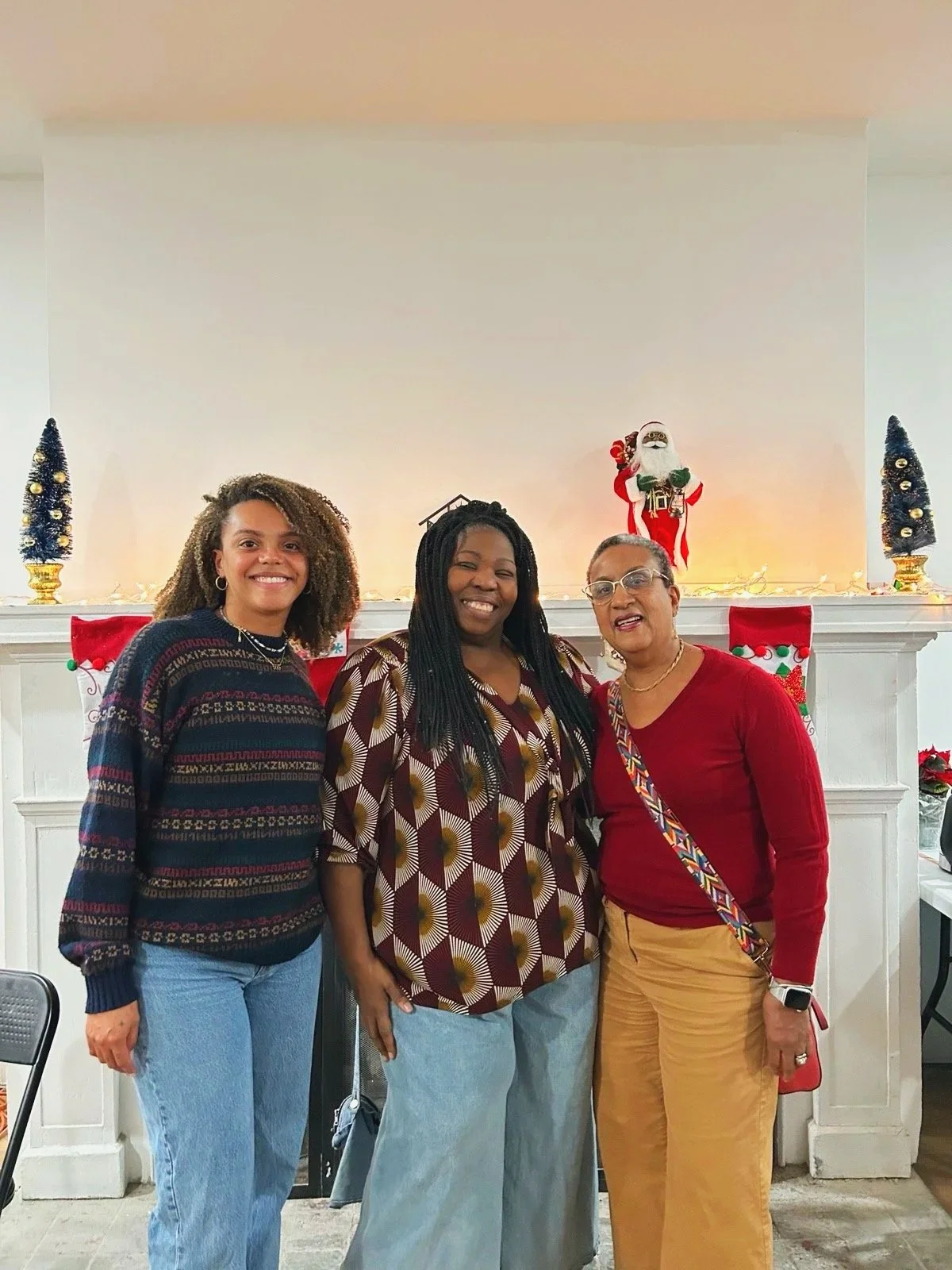 Three women standing in front of a fireplace decorated with Christmas stockings, a Santa figurine, and holiday decor, smiling at the camera.