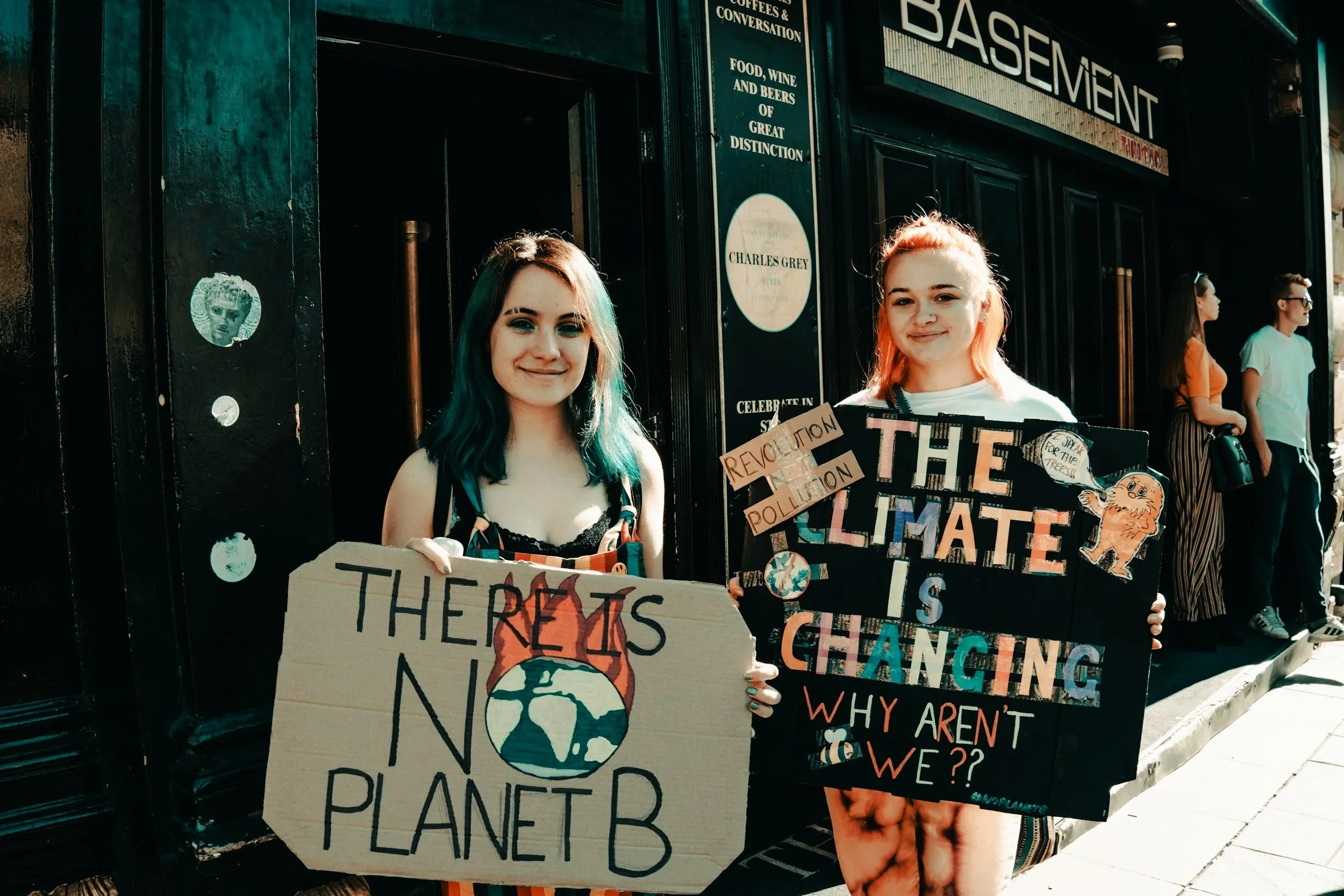 Two young women standing outside holding protest signs about climate change. The woman on the left holds a sign that reads 'There is no Planet B' with an illustration of the Earth. The woman on the right holds a sign that says 'The Climate is Changing, Why Aren't We??' and has smaller notes that mention revolution and pollution.