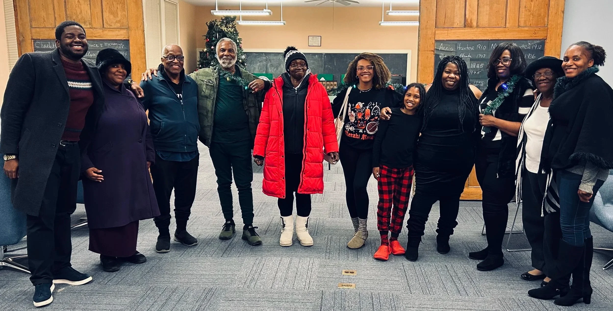 Group of twelve diverse people standing together indoors, smiling, with Christmas decorations and a chalkboard in the background.