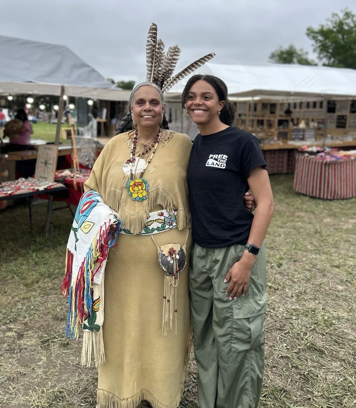 Two women standing together at an outdoor market, with booths behind them. The woman on the left wears traditional indigenous clothing with feathers and colorful accessories, while the woman on the right wears a black T-shirt that says 'Free the Land' and green pants. They are smiling and posing for the photo.