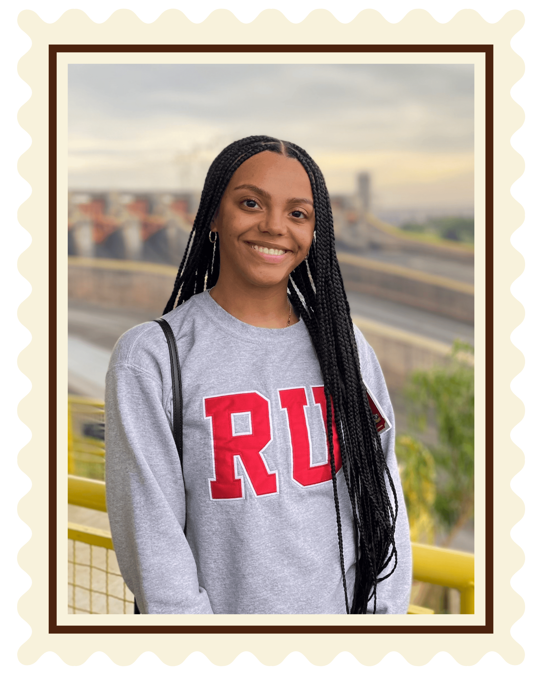 A young woman with long braids smiling outdoors on a bridge, wearing a gray sweatshirt with red 'RU' letters, black strap purse, and earrings, with a bridge and city in the background.