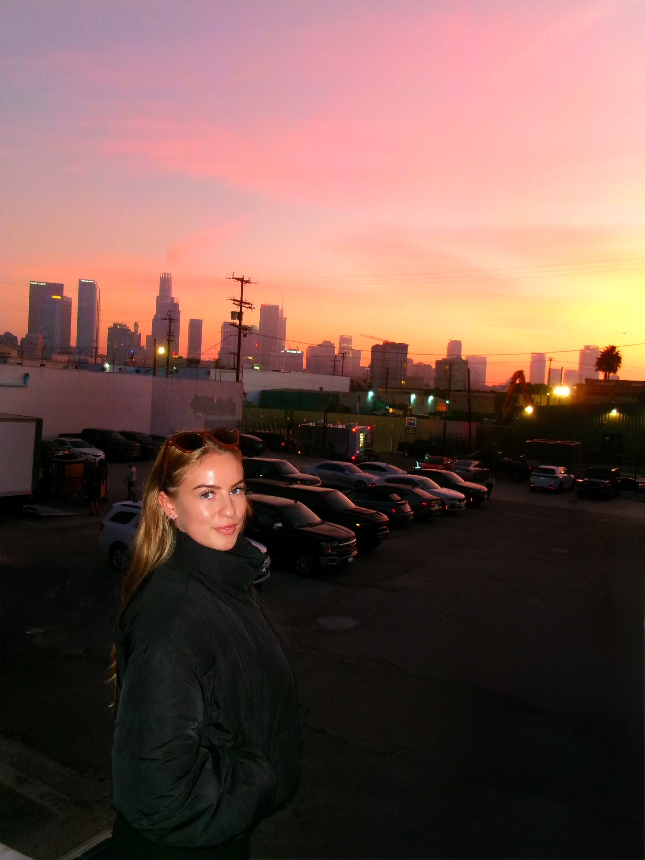 A woman with blonde hair wearing a black jacket stands in a parking lot at sunset, with the Los Angeles skyline in the background.