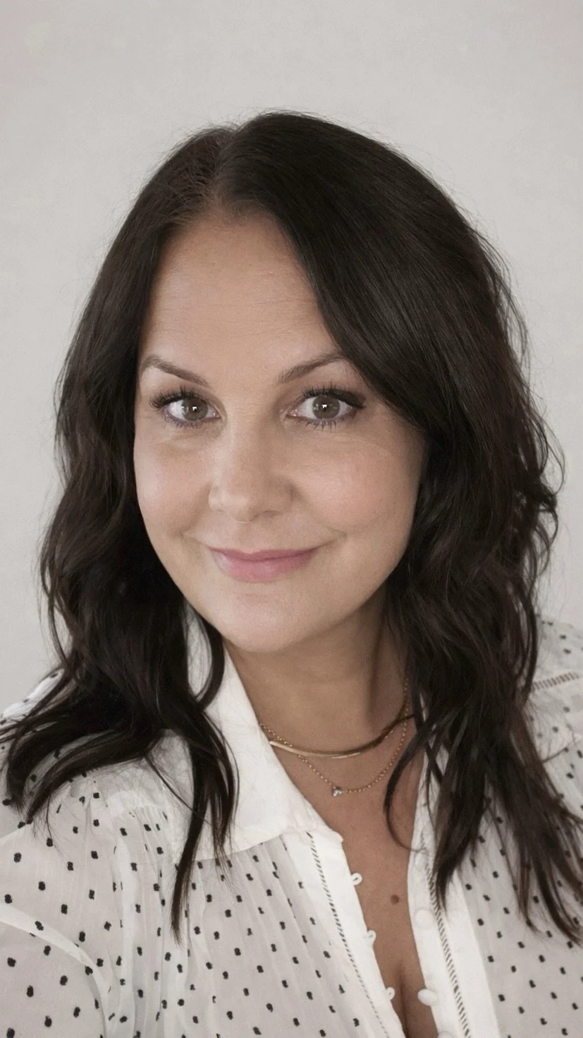 A woman with shoulder-length dark wavy hair, light skin, and gray eyes, smiling at the camera, wearing a white shirt with black polka dots and layered gold necklaces.