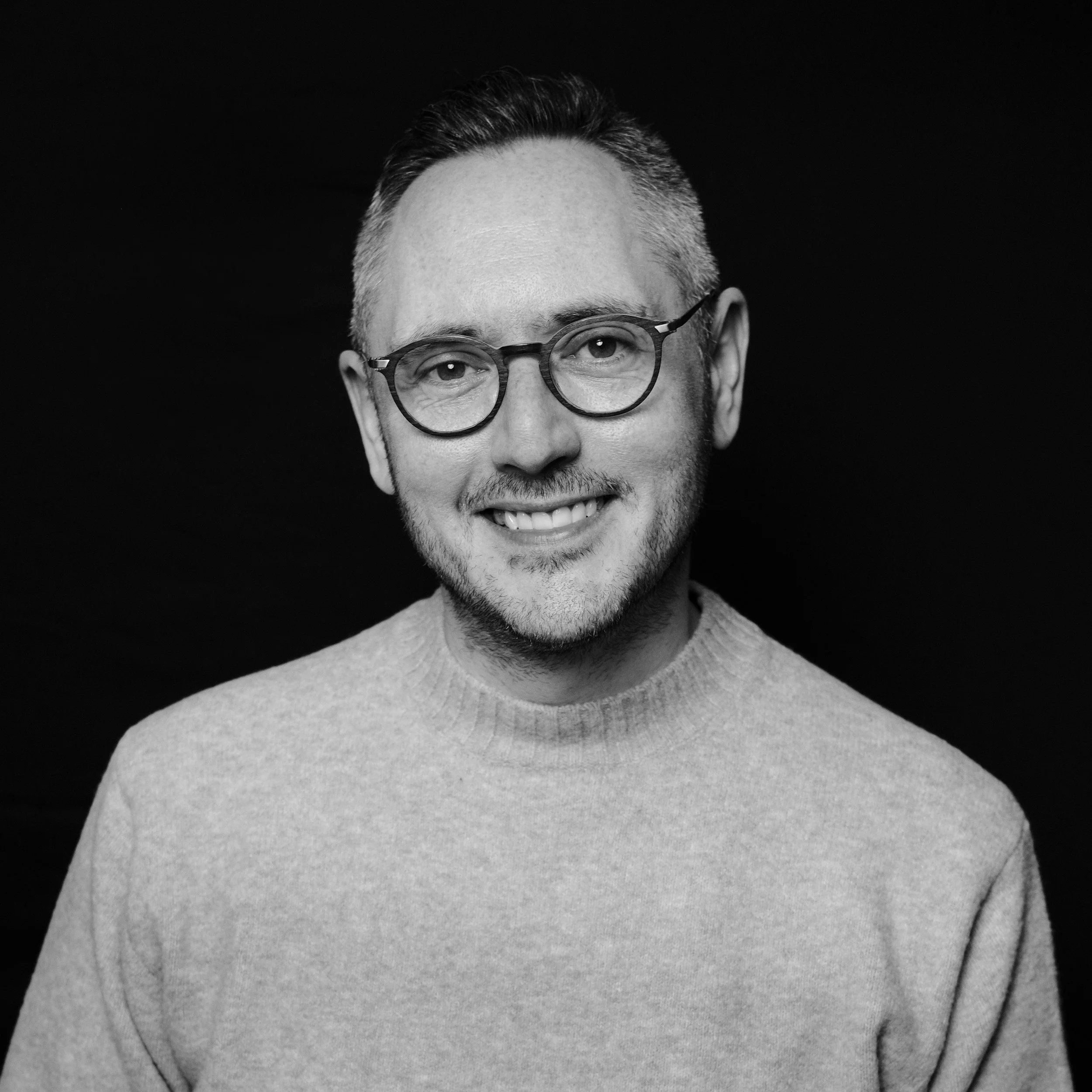 Close-up black and white photo of a smiling man wearing glasses, with short hair, a beard, and a knit sweater, against a dark background.