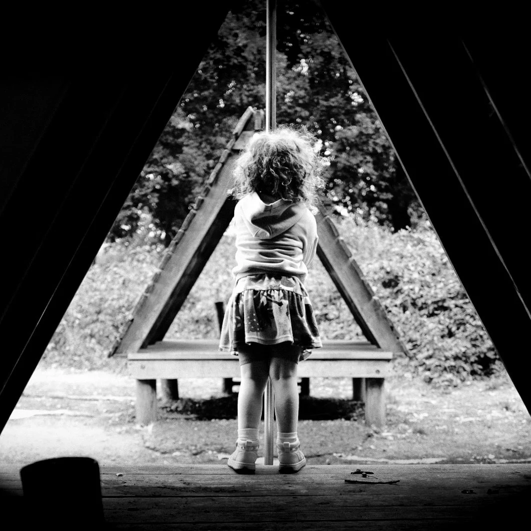 A young girl with curly hair standing on a wooden platform, facing away, inside a teepee-shaped structure, looking out at a wooden triangular play feature outdoors with trees in the background in black and white.