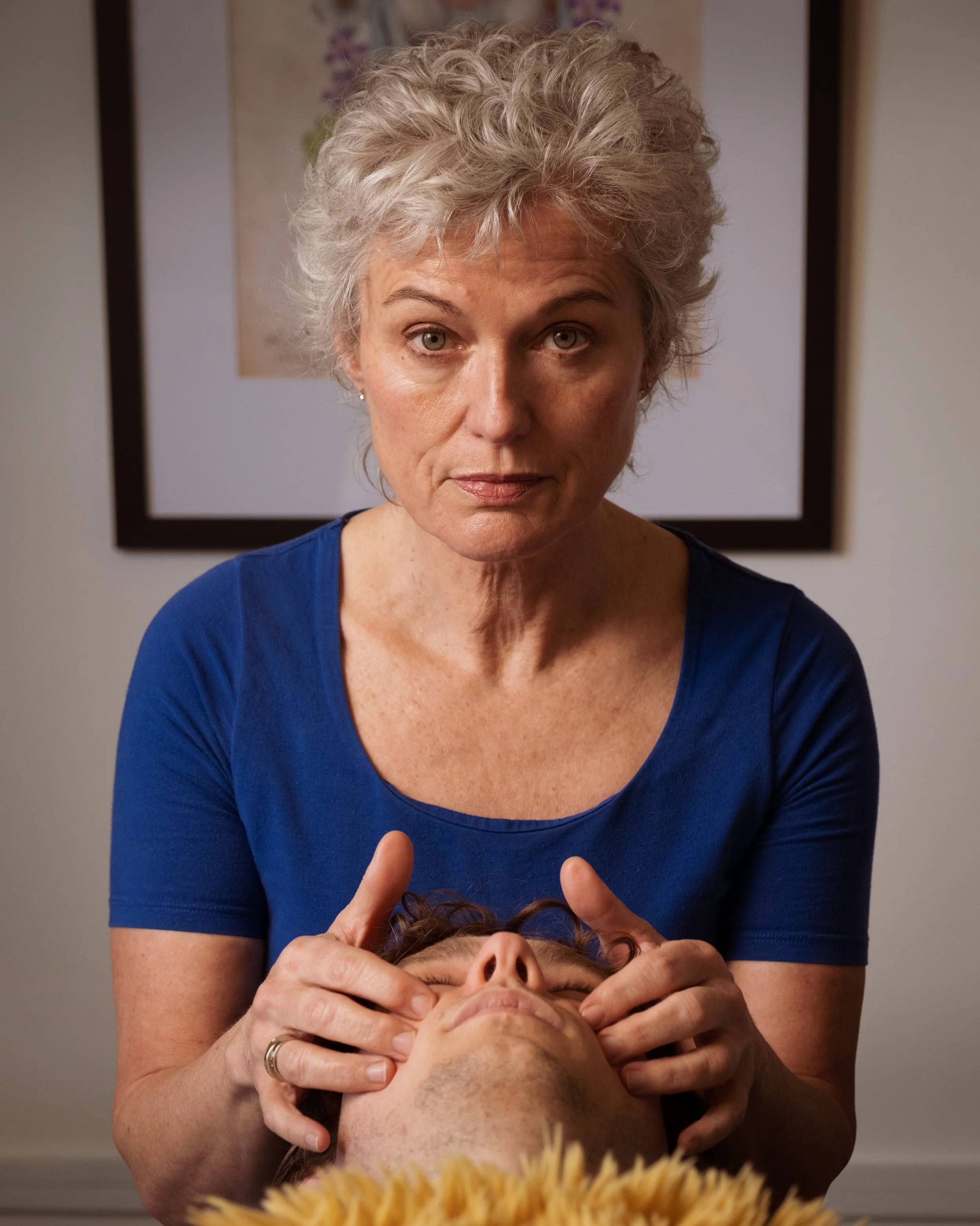 A woman with short curly gray hair giving a massage or facial to a person lying down with their eyes closed. The woman is wearing a blue top and is holding the person's head gently. The background shows a framed picture on the wall.