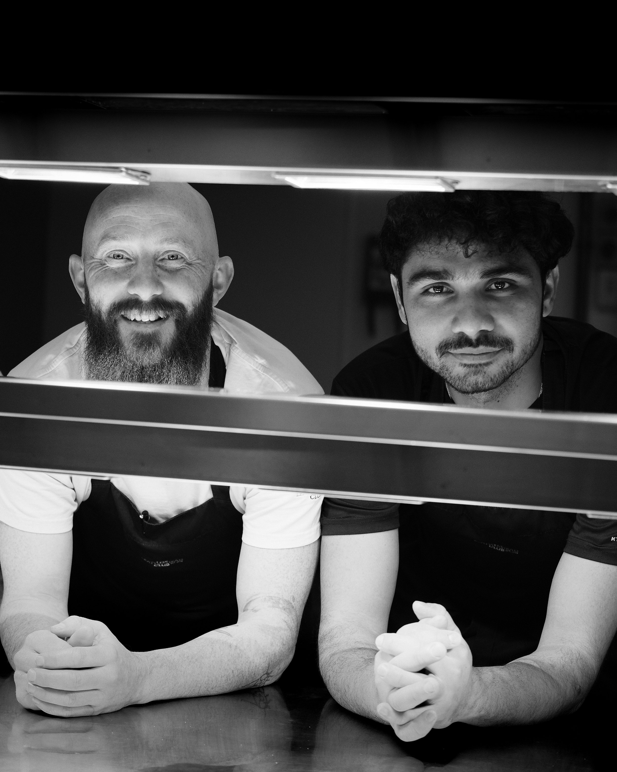 Two men sitting at a table with their arms crossed and hands clasped in front of a black background, seen from below through a grate or vent.