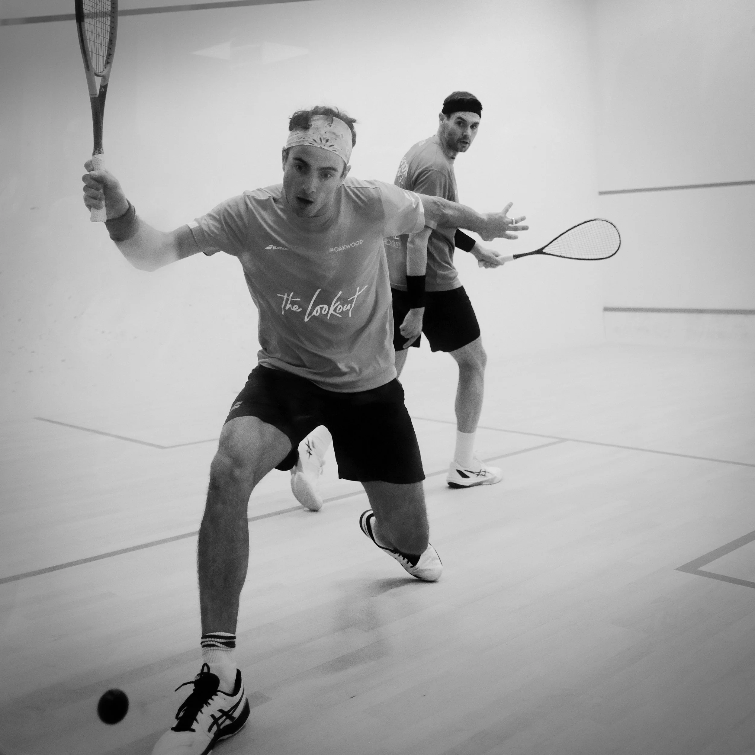 Two men playing squash on an indoor court, one reaching out to hit a ball with a racket, both wearing sportswear and headbands.