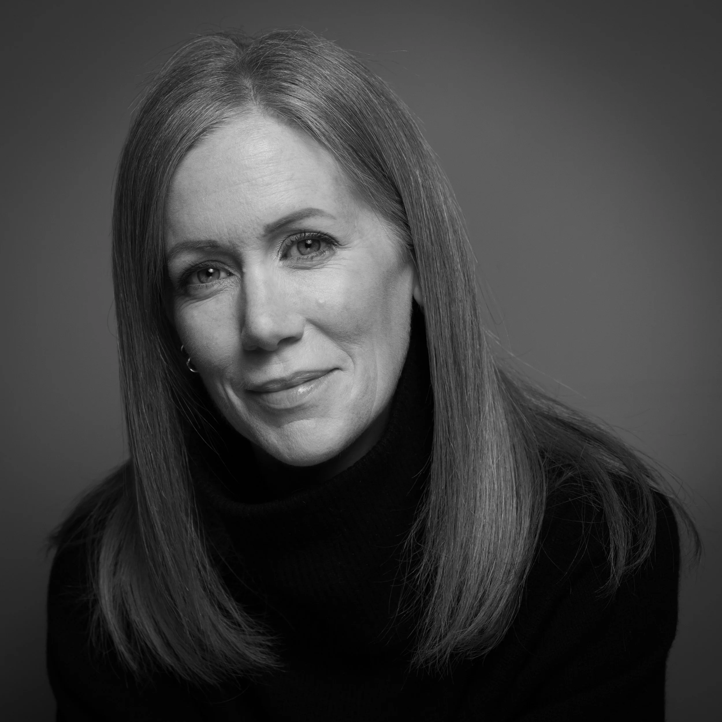 Black and white close-up of a woman with straight hair, posing in front of a plain background.