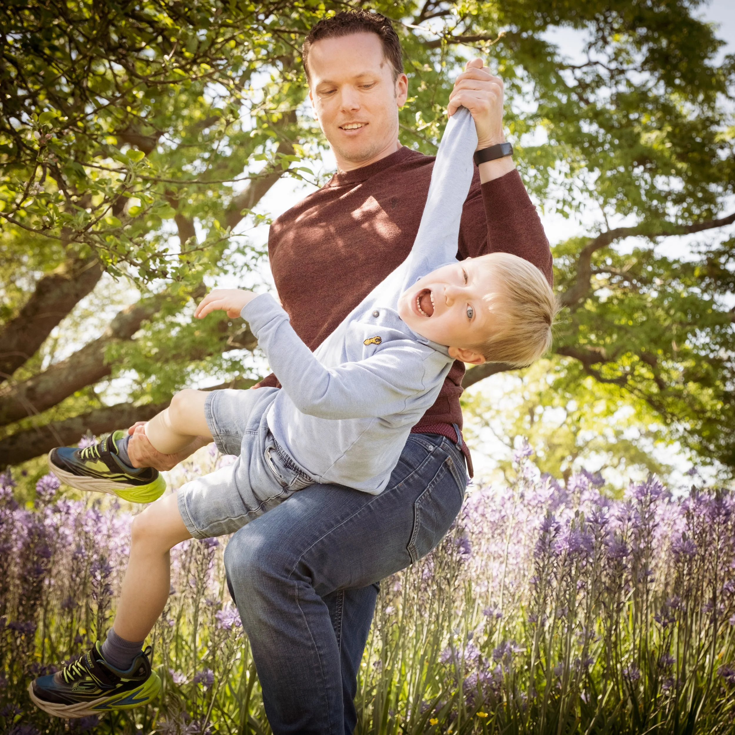 A man holding a laughing child on a swing in a park with trees and purple flowers in the background.