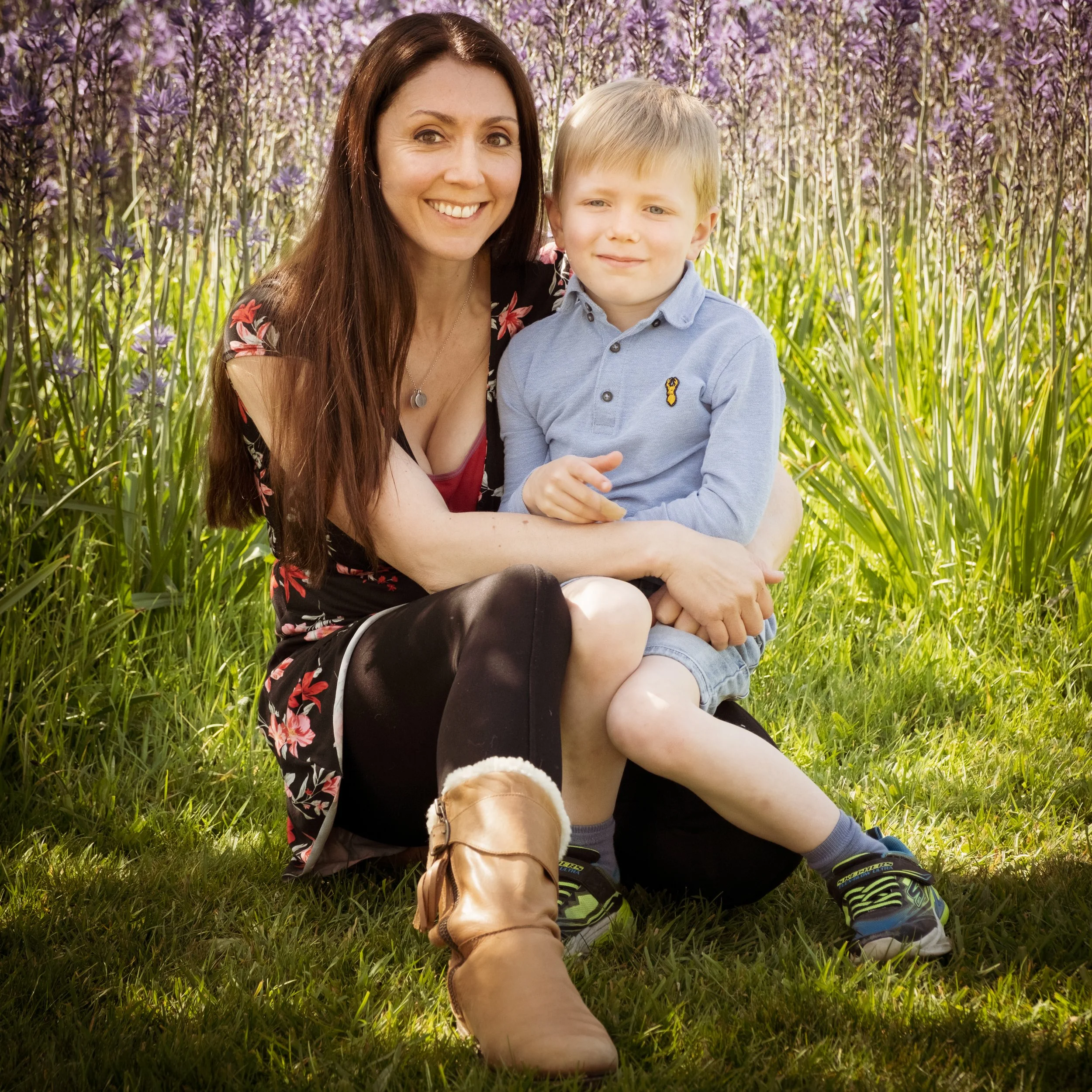 A woman with long brown hair and a boy with short blonde hair sitting in a field of purple flowers, smiling at the camera. The woman is wearing a black dress with a floral pattern, and the boy is wearing a light blue button-up shirt.