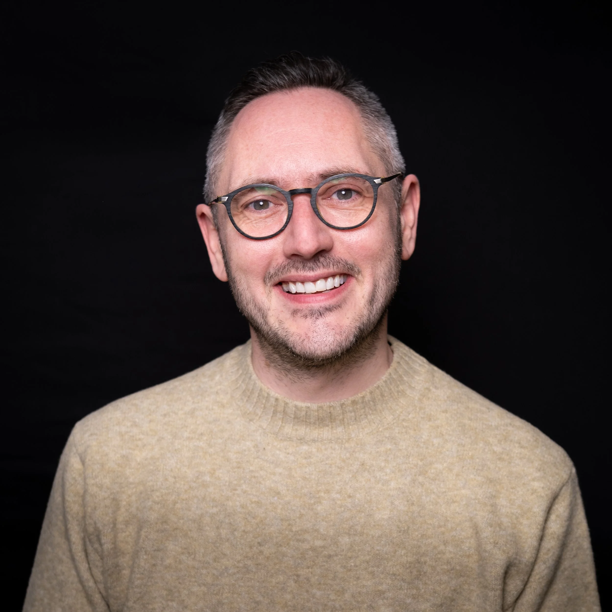 A close-up of a smiling man with short dark hair, wearing round glasses with black rims and a beige sweater, against a black background.