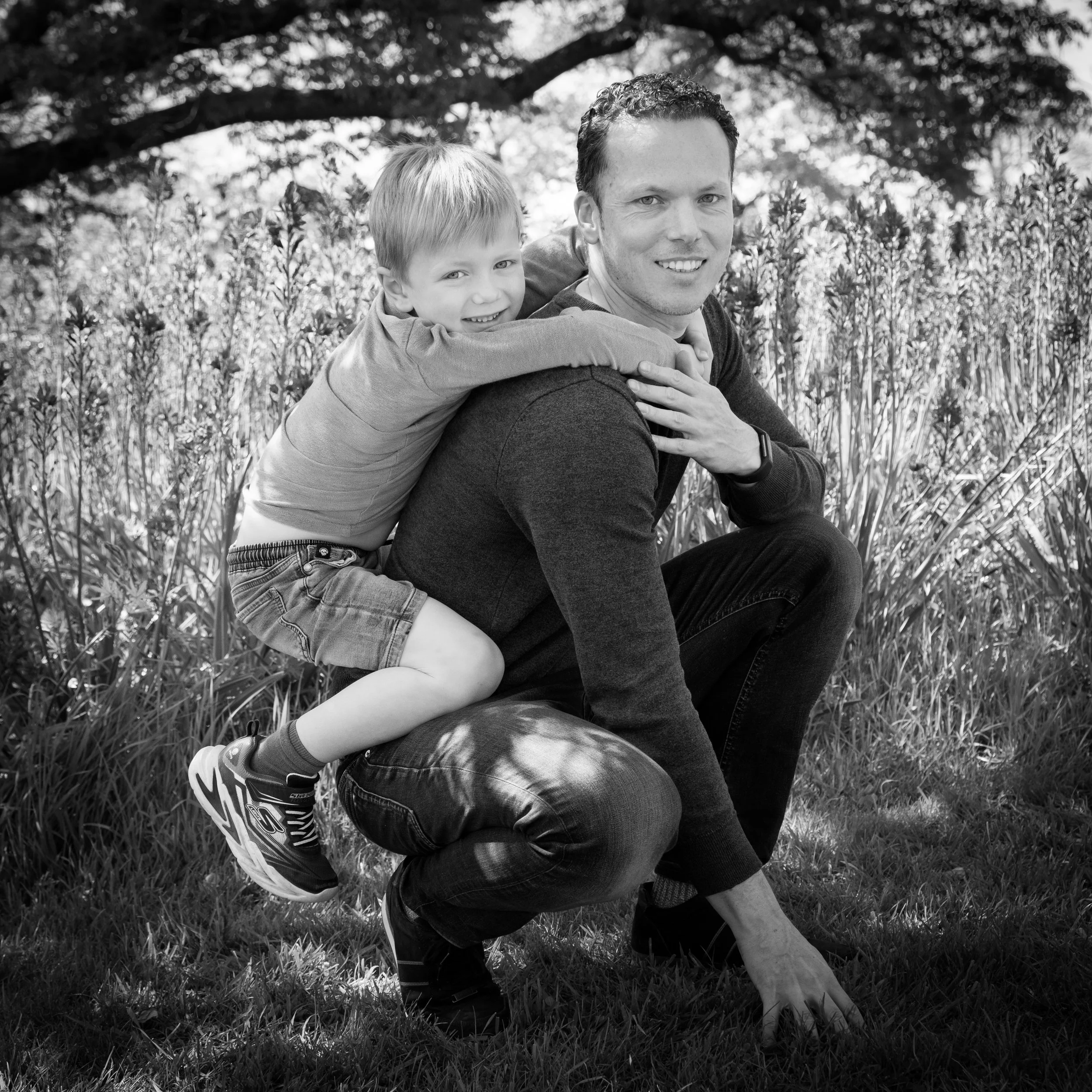 A man and a young boy are outdoors in a grassy area with trees and bushes in the background. The boy is hitching a ride on the man's back, smiling, while the man is crouched down and smiling at the camera.