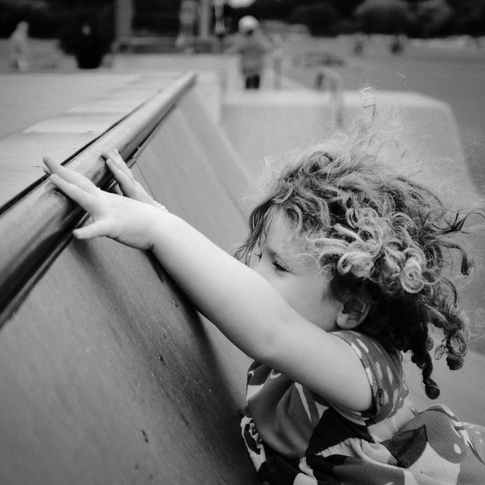 A young child with curly hair leaning on a railing, reaching out and looking intently at something below or beyond it.