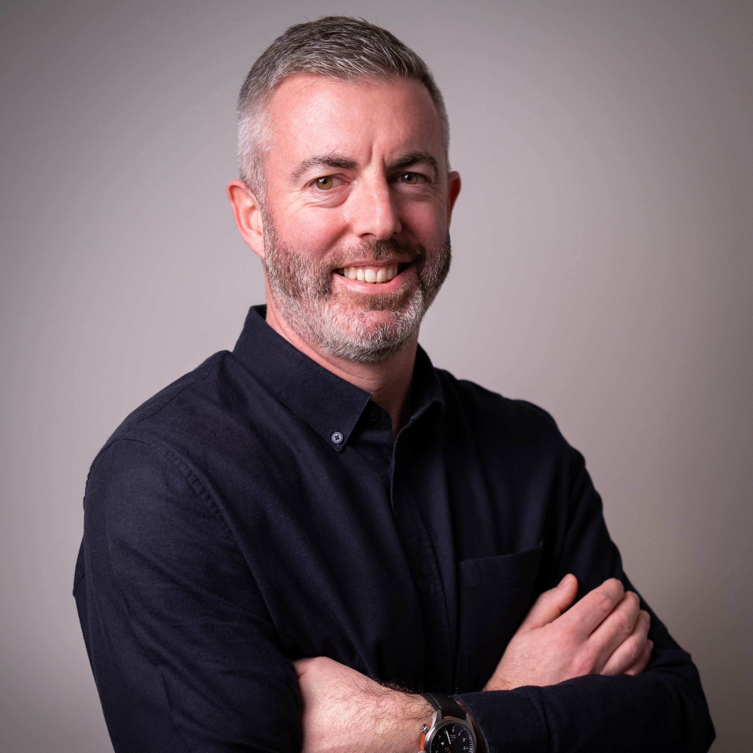 A smiling man with short gray hair and beard, wearing a dark shirt and watch, with arms crossed, against a plain gray background.