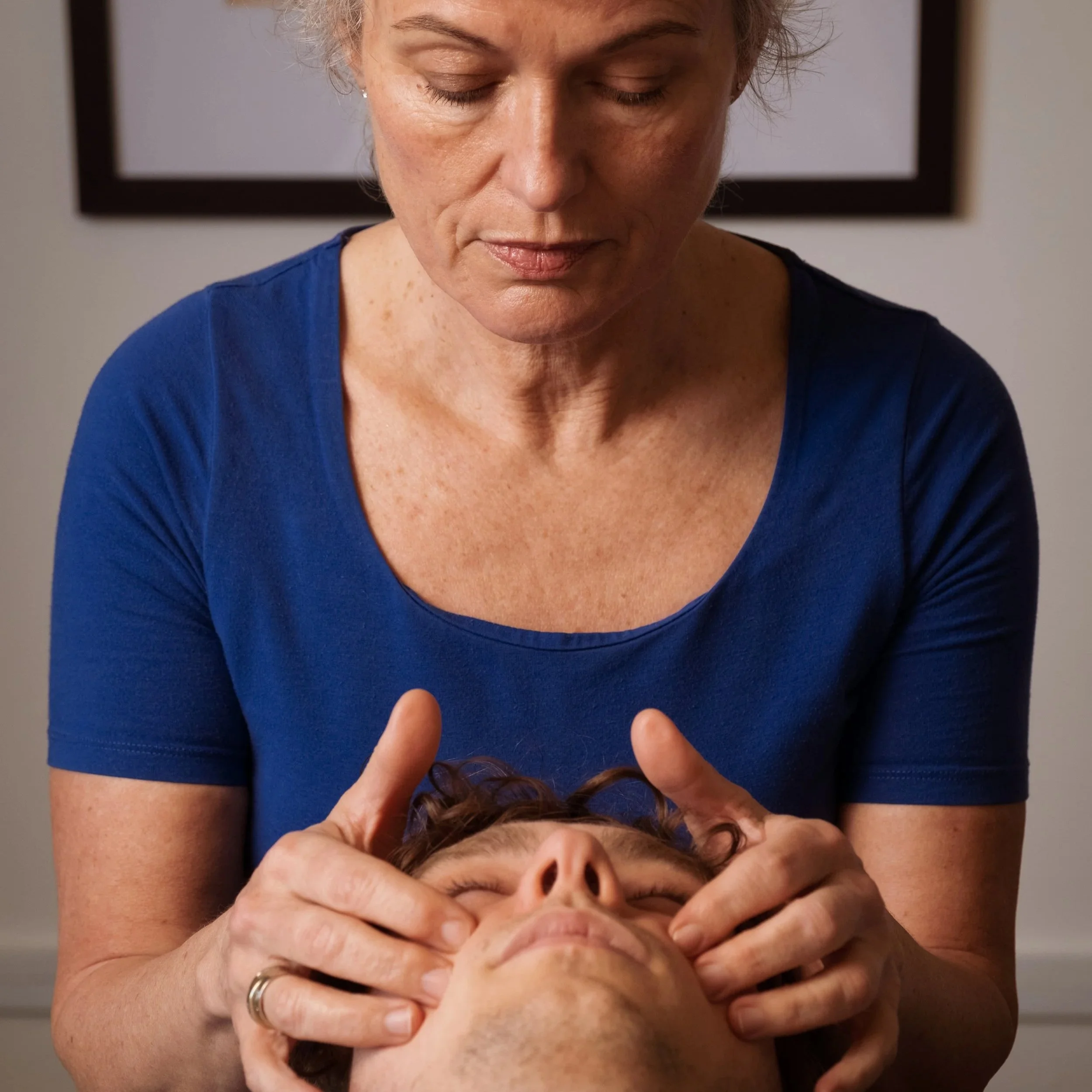 A woman with closed eyes and a neutral expression is holding a person's head in her hands, with the person's face partially visible. The woman is wearing a blue shirt and appears to be inspecting or comforting the person.