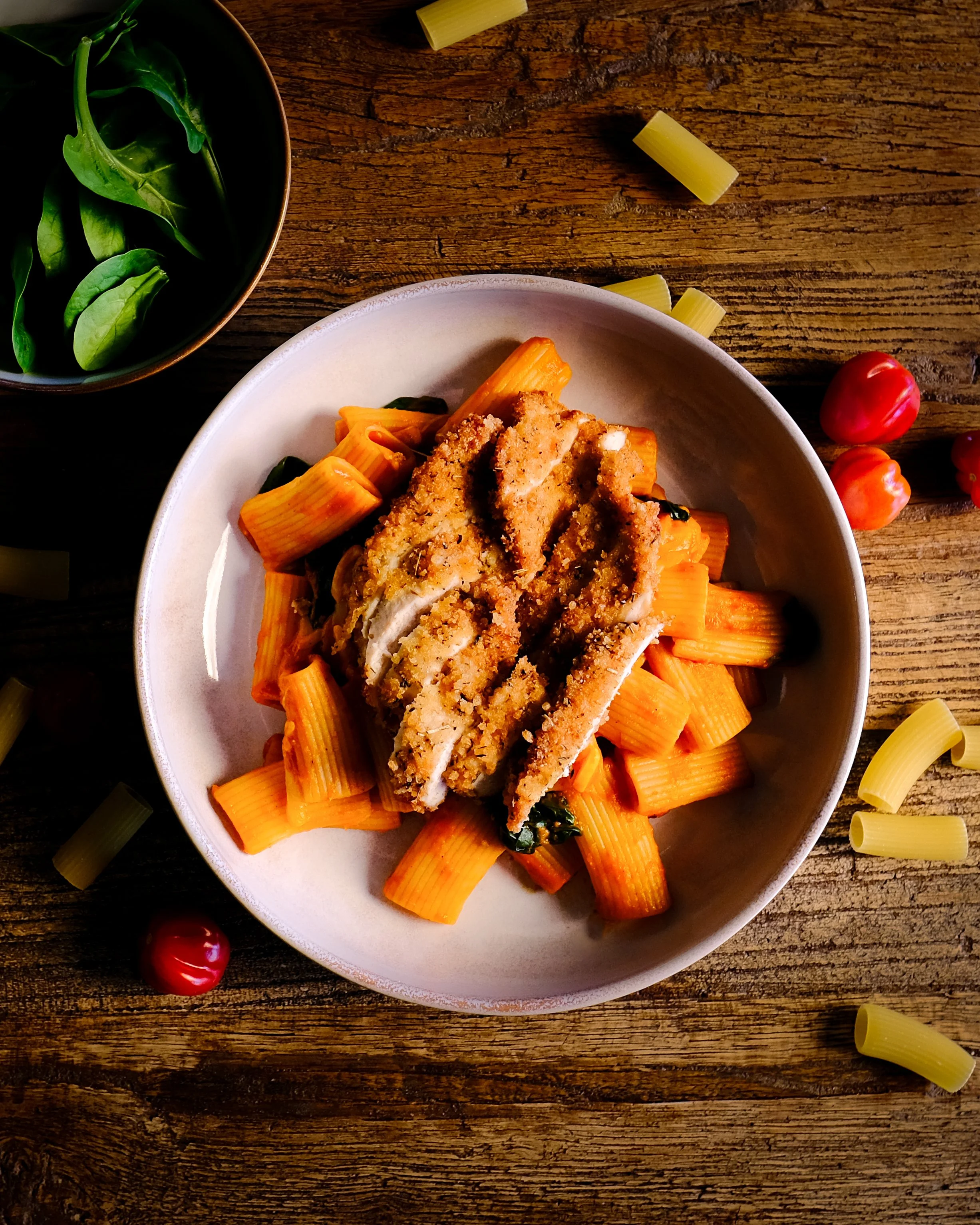 A top-down view of a plate with baked chicken covered in breadcrumb topping, served over cooked rigatoni pasta with spinach, on a rustic wooden table. Nearby are cherry tomatoes, uncooked rigatoni pasta, and a small bowl of fresh basil leaves.