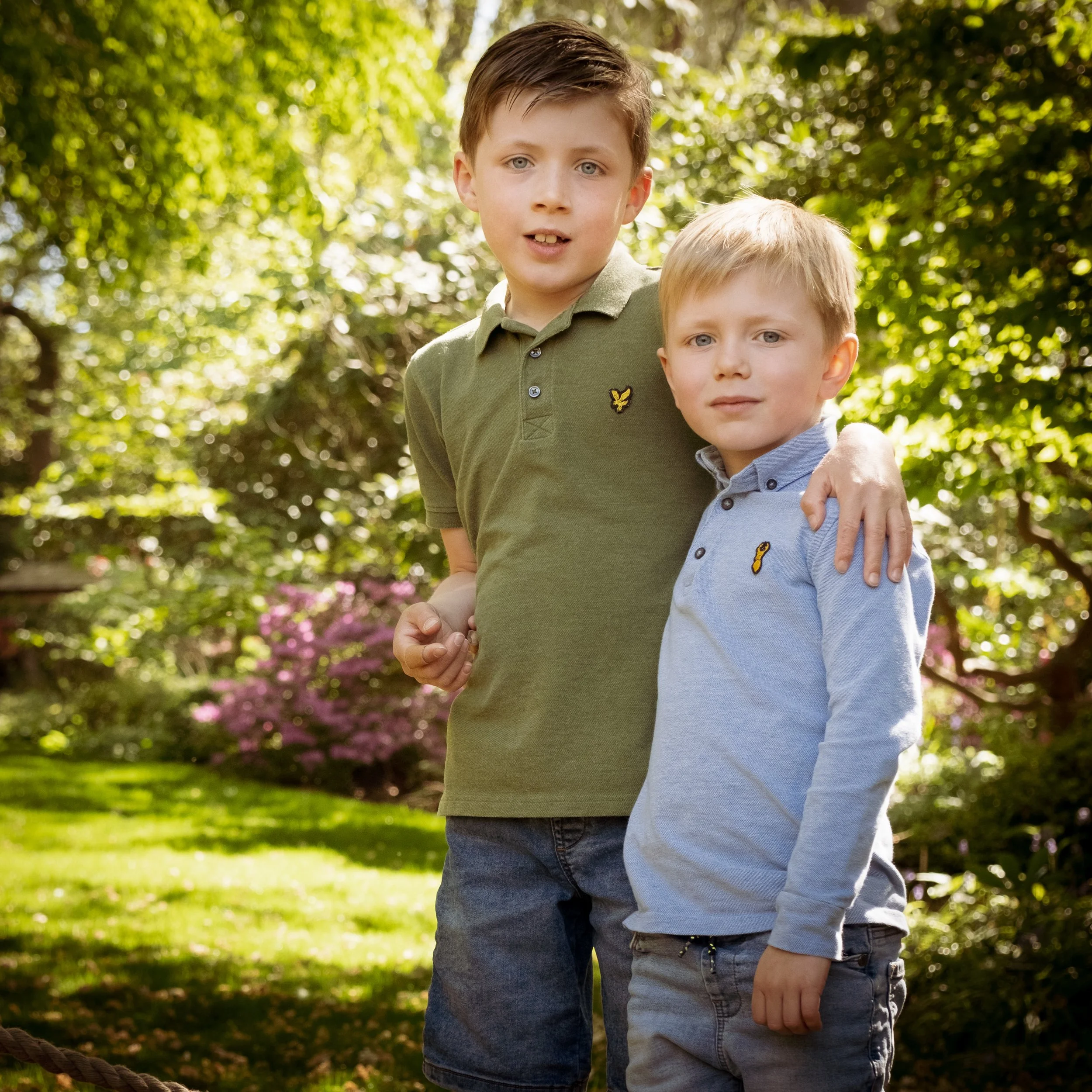 Two young boys standing outdoors in a garden or park area with green trees and bushes, one with brown hair wearing a green shirt and the other with light brown hair wearing a light blue shirt.