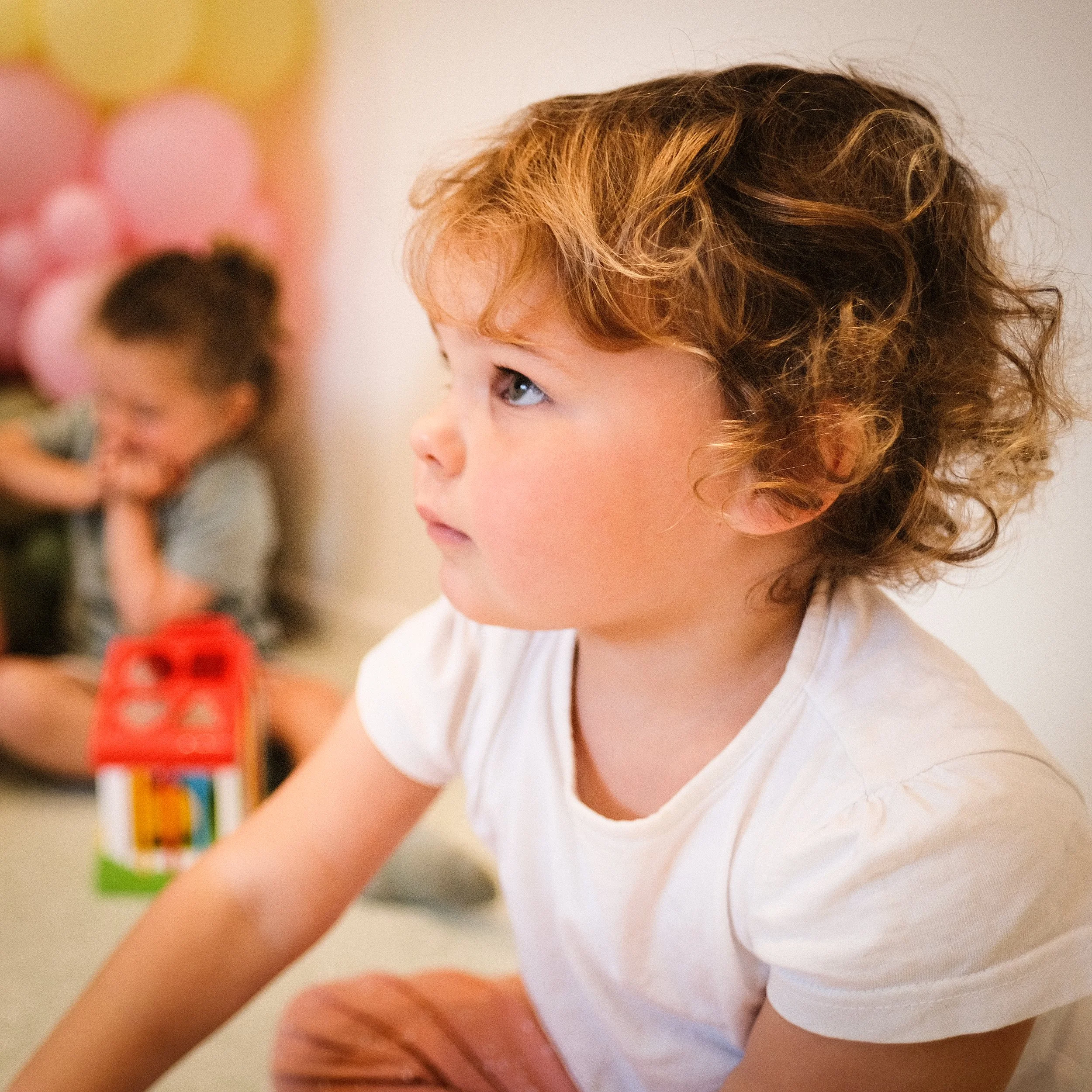 A young child with curly brown hair and blue eyes, wearing a white shirt, looking to the side in an indoor setting.