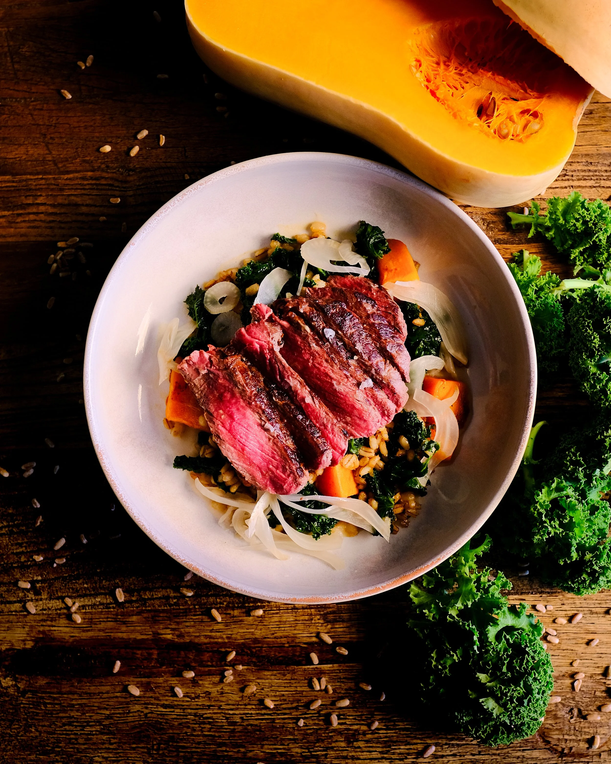 A bowl of cooked grains topped with sliced steak, vegetables, and garnished with sliced onions. Behind the bowl, there is a large piece of yellow squash and some fresh curly kale on a rustic wooden surface with scattered grains.