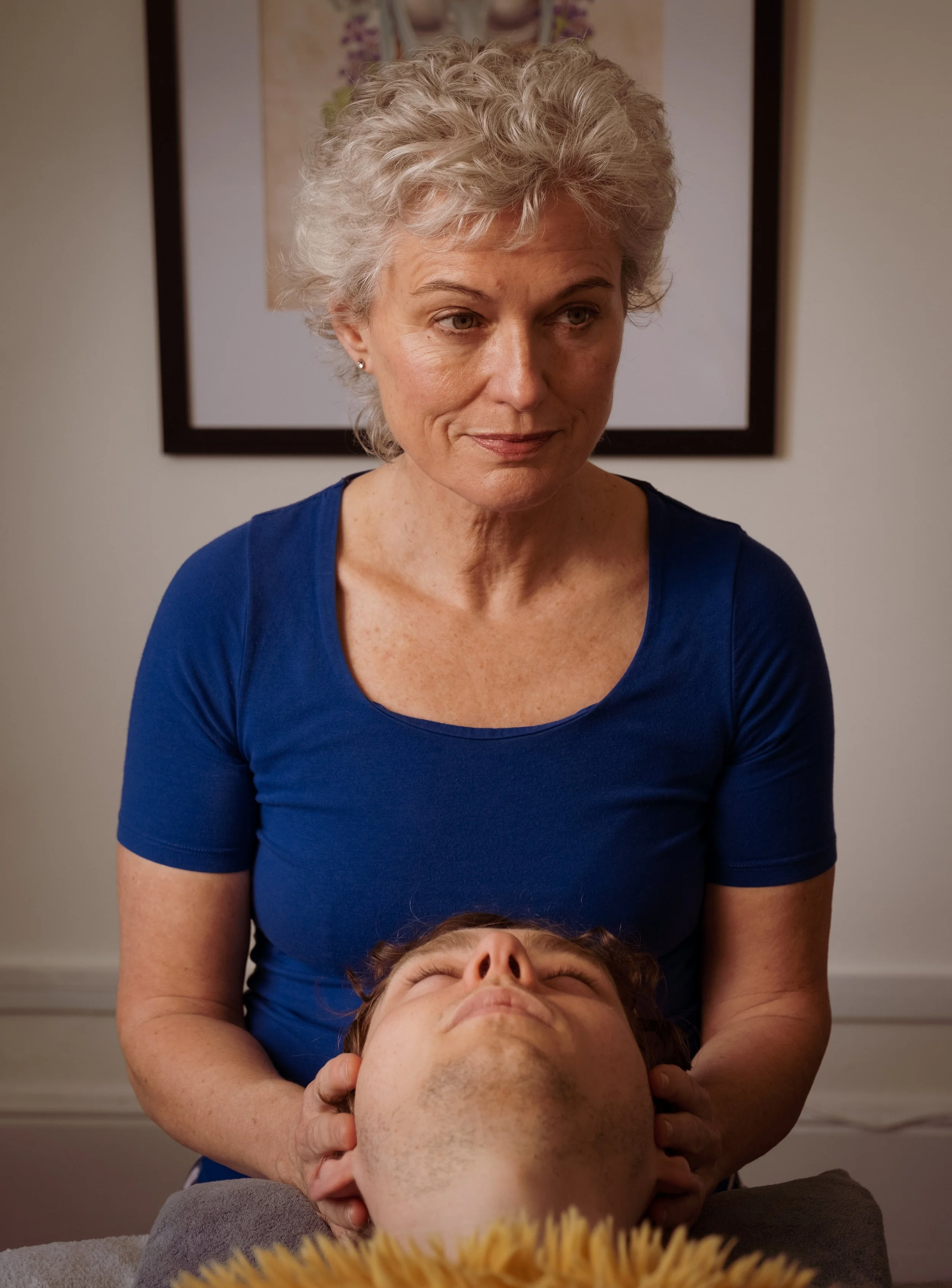 A woman providing a neck massage to a person lying down with their eyes closed, in a room with a framed floral artwork on the wall.