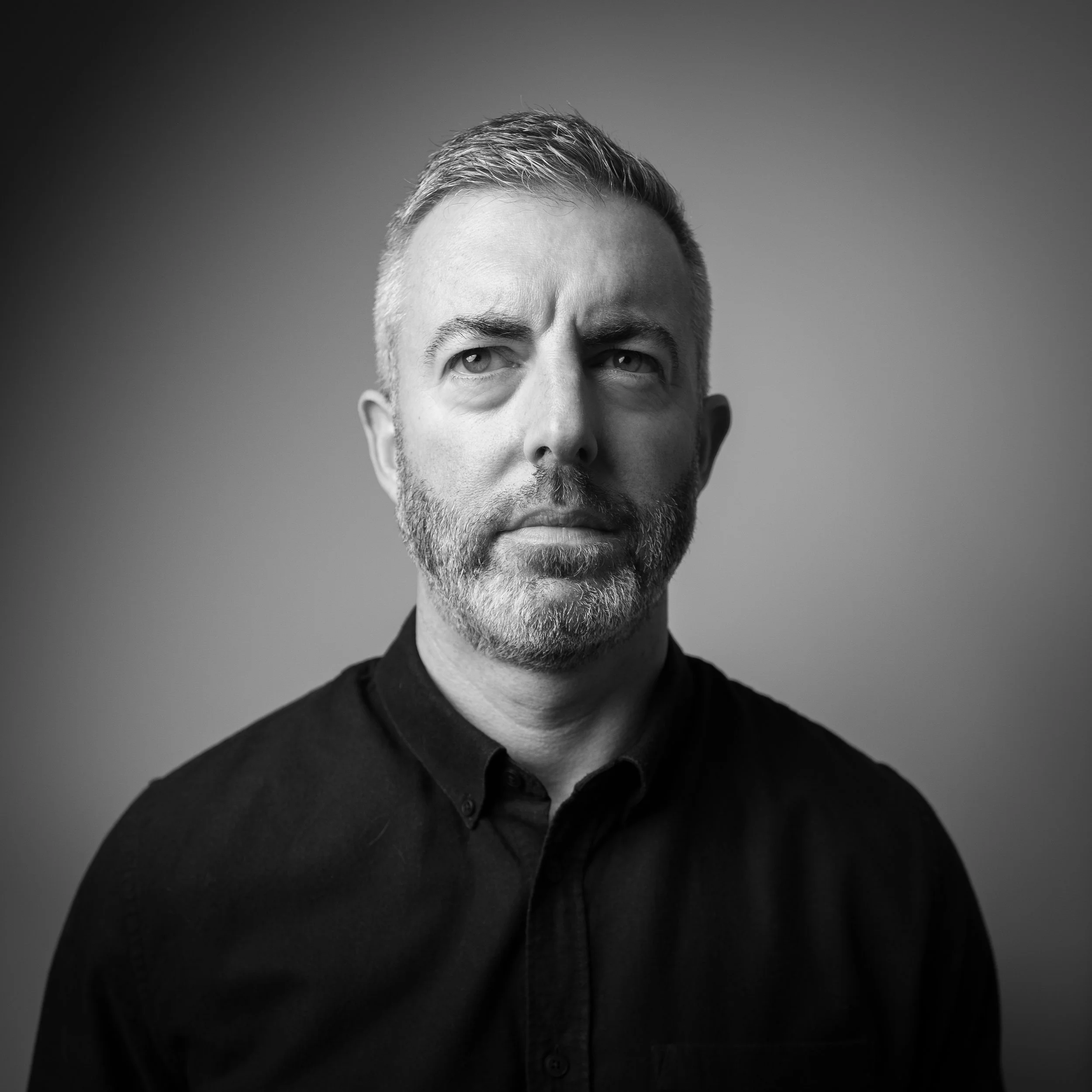 Black and white portrait of a middle-aged man with short hair and a beard, wearing a dark button-up shirt, against a plain gray background.