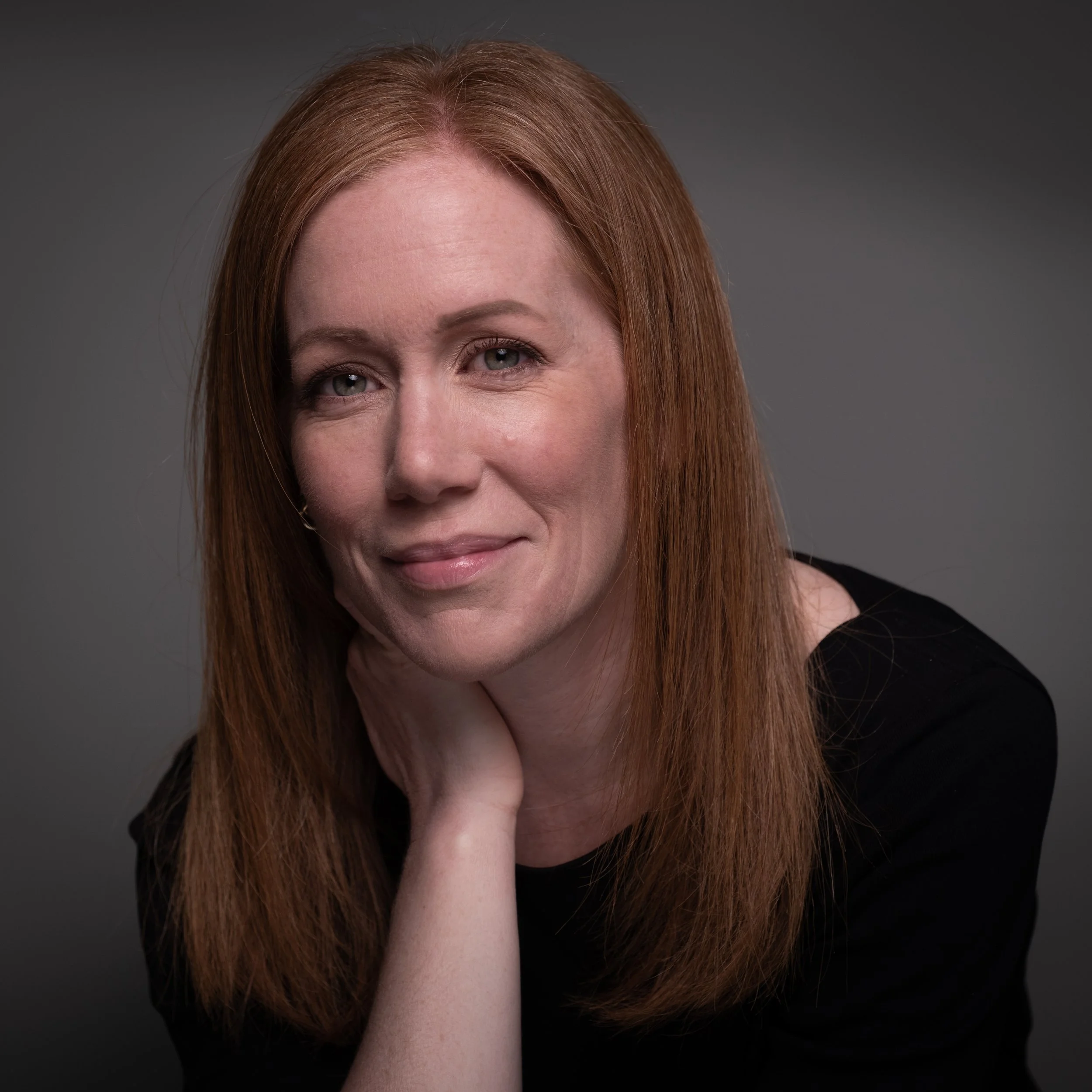 Close-up of a woman's face with ginger hair, green eyes, and light skin, resting her hand on her neck against a dark gray background.