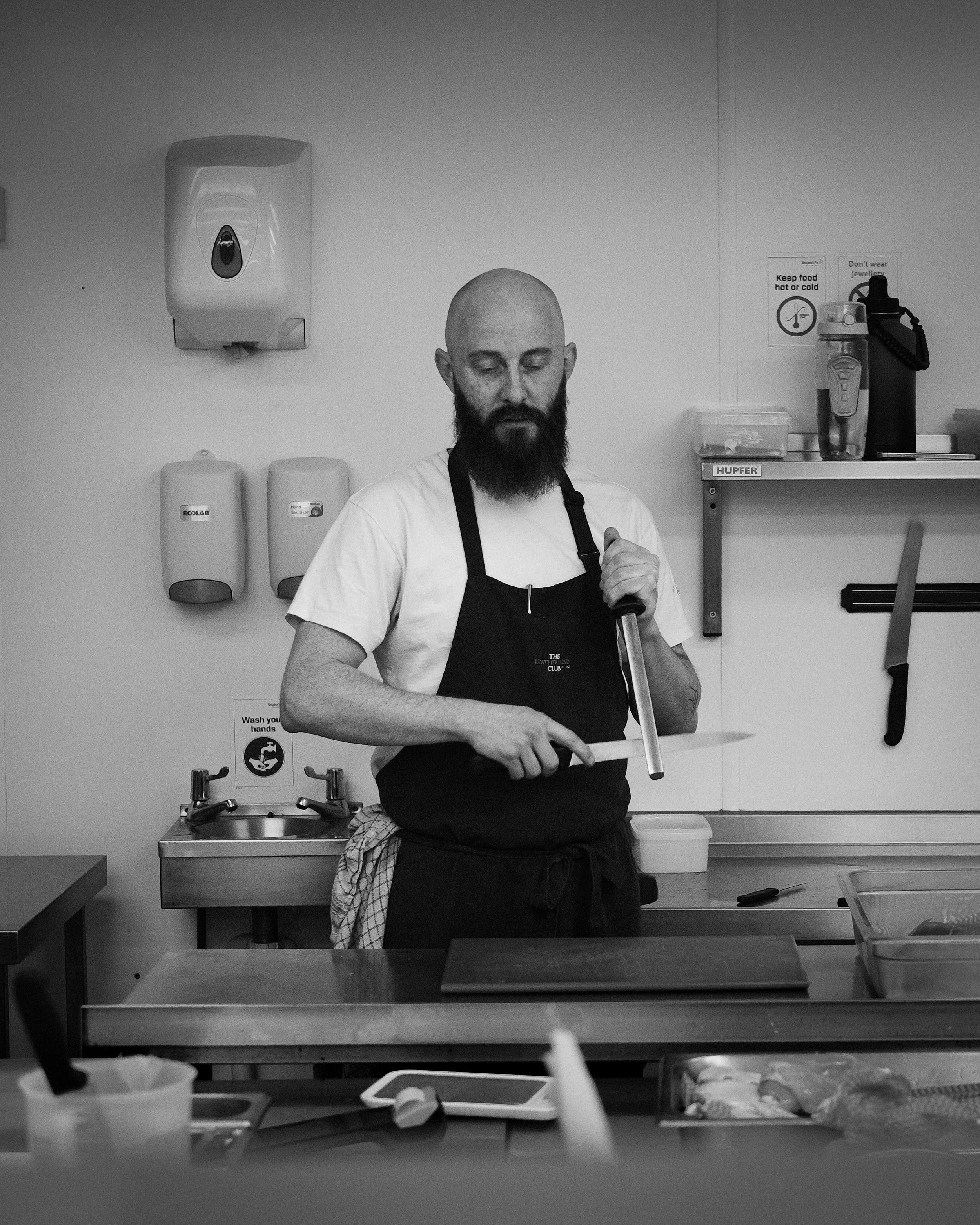 A bald man with a beard wearing a white shirt and black apron, standing in a kitchen, holding a kitchen utensil. The kitchen has soap dispensers on the wall, and a shelf with a box labeled "HUPFER" and a container. There are signs on the wall with instructions about food safety and jewelry, and a handwashing sign.