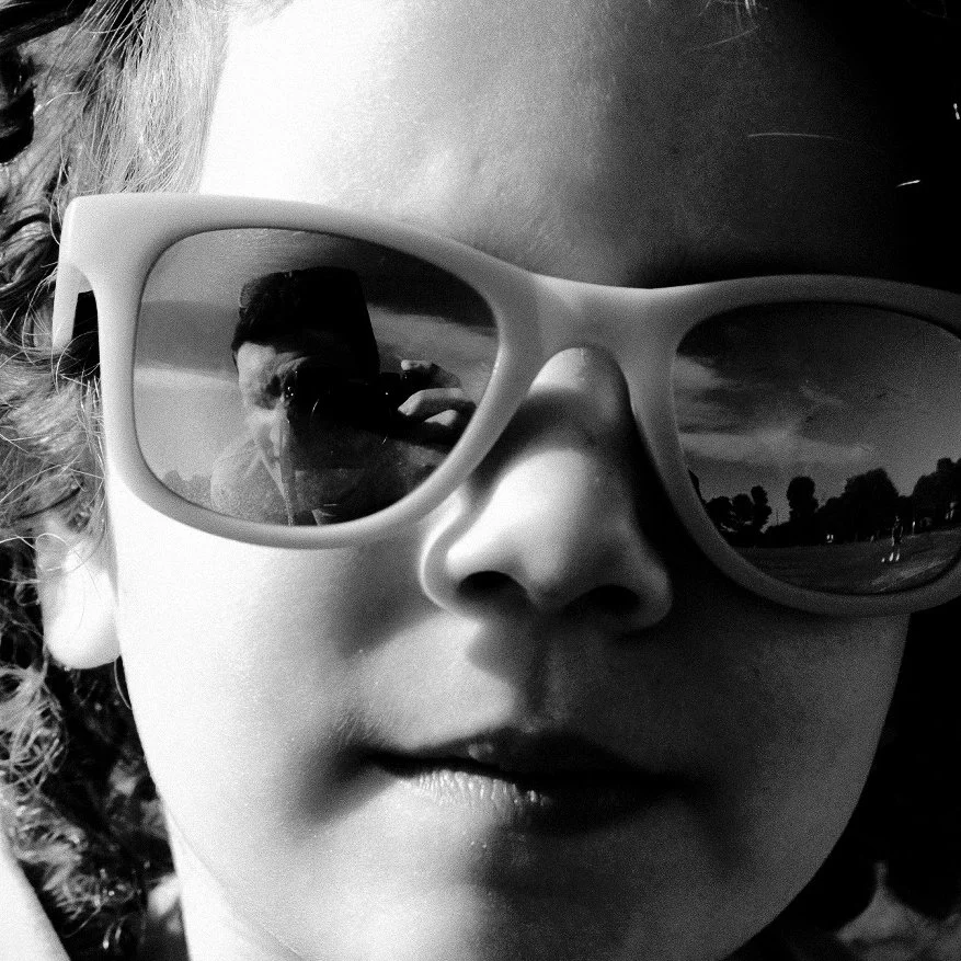 Close-up of a young girl wearing large sunglasses, reflecting a person taking her photo and an outdoor scene with trees and a cloudy sky.