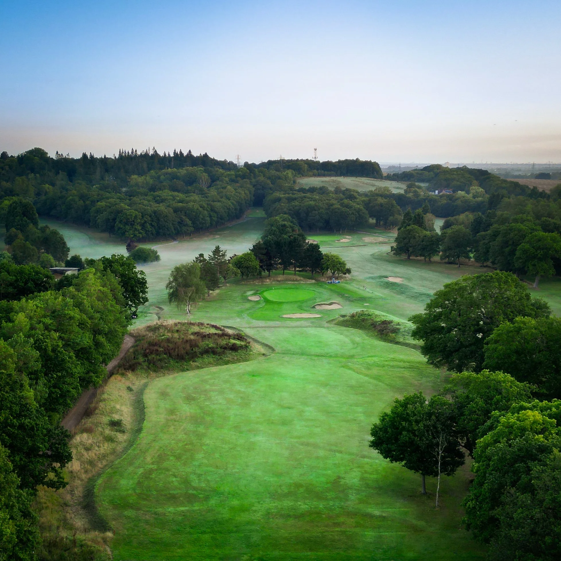 A scenic view of a golf course with green fairways and sand bunkers, surrounded by lush trees and rolling hills, under a clear sky.