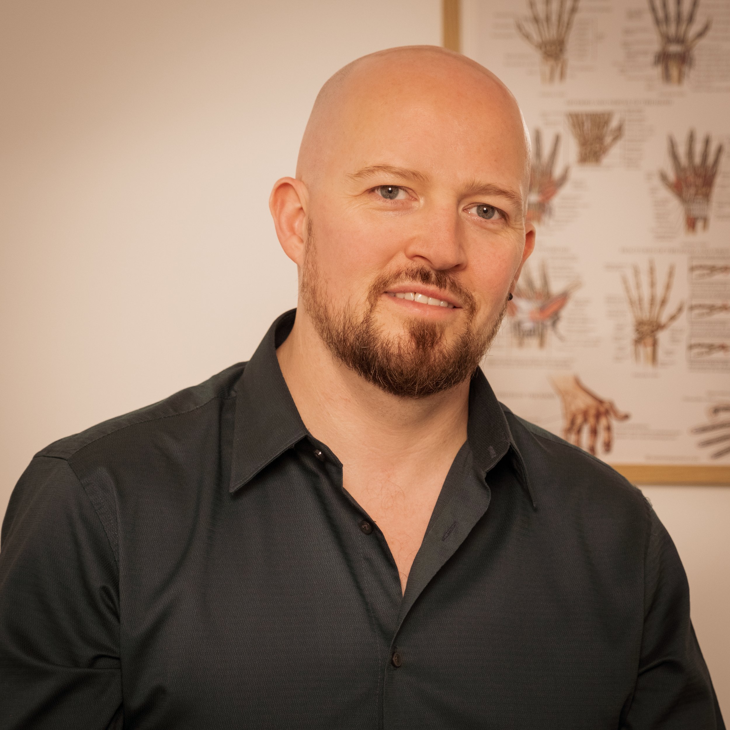 A man with a shaved head and a beard, smiling, wearing a black shirt, standing indoors with a poster of scientific diagrams in the background.