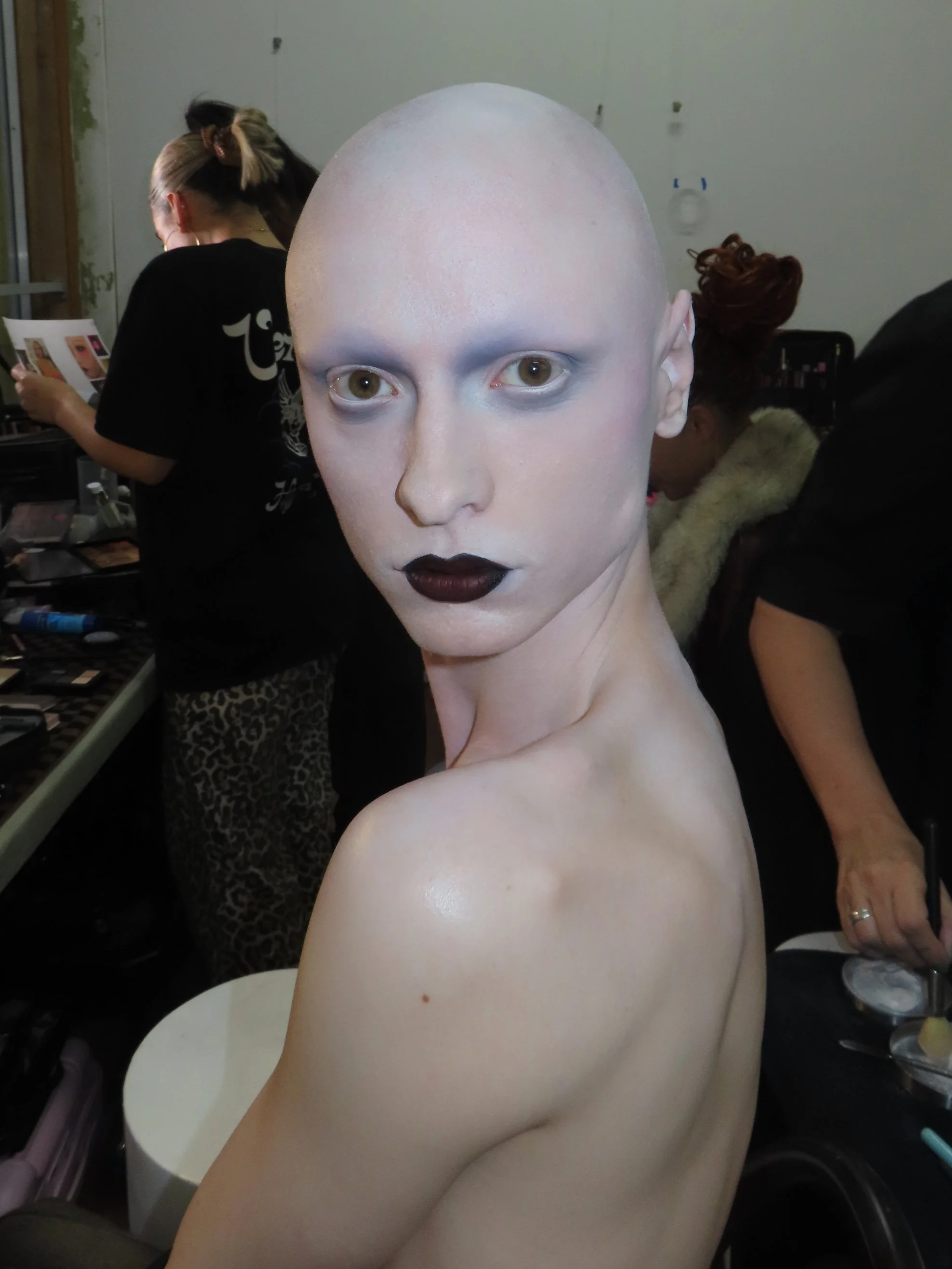 A mannequin with painted white skin, black lipstick, and no hair, seated in a room with people preparing makeup in the background.