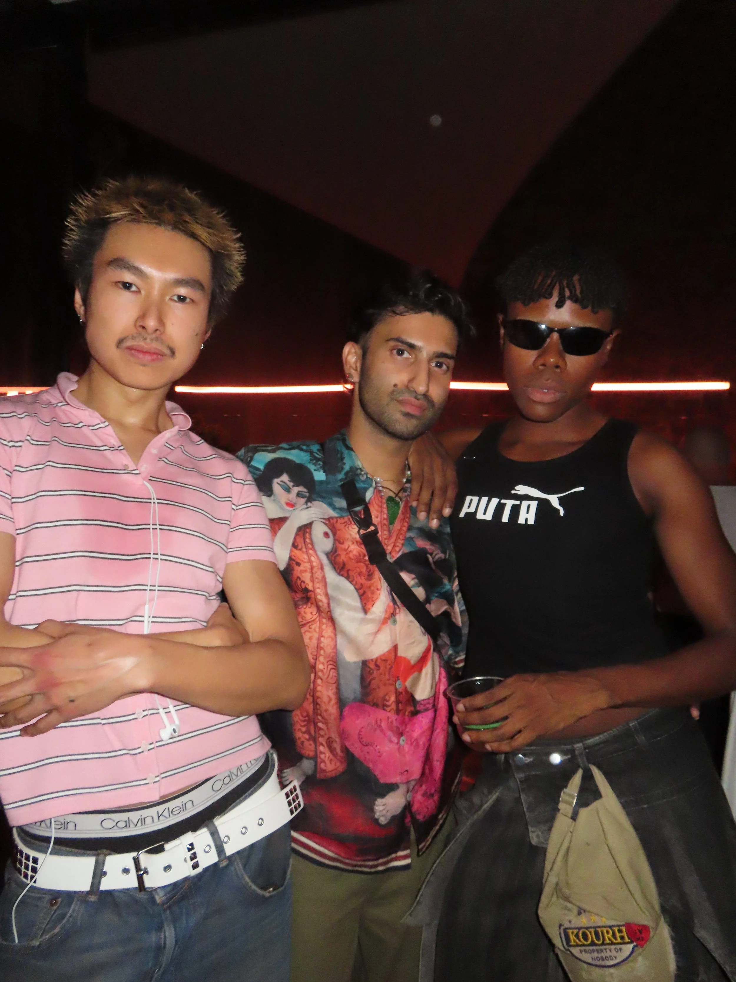 Three young men in a dark room with red lighting, standing close together and posing for a photo. The individual on the left is wearing a pink striped shirt, the middle person is wearing a colorful shirt with a woman design, and the person on the rig