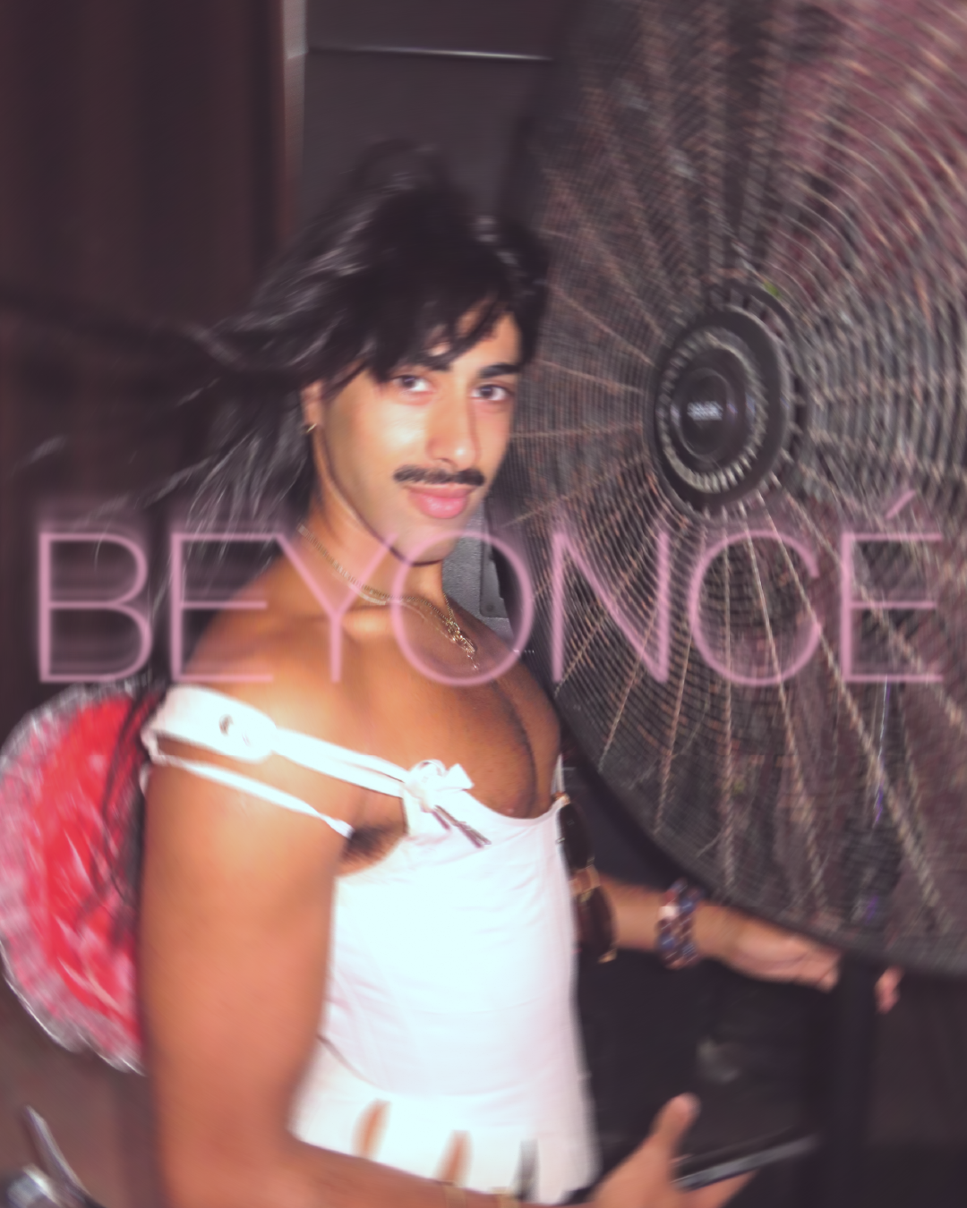 A person with long dark hair, a mustache, and a slight smile, wearing a white dress with shoulder straps and holding a phone, standing beside a large fan with the word 'BEYONCE' in pink neon letters.