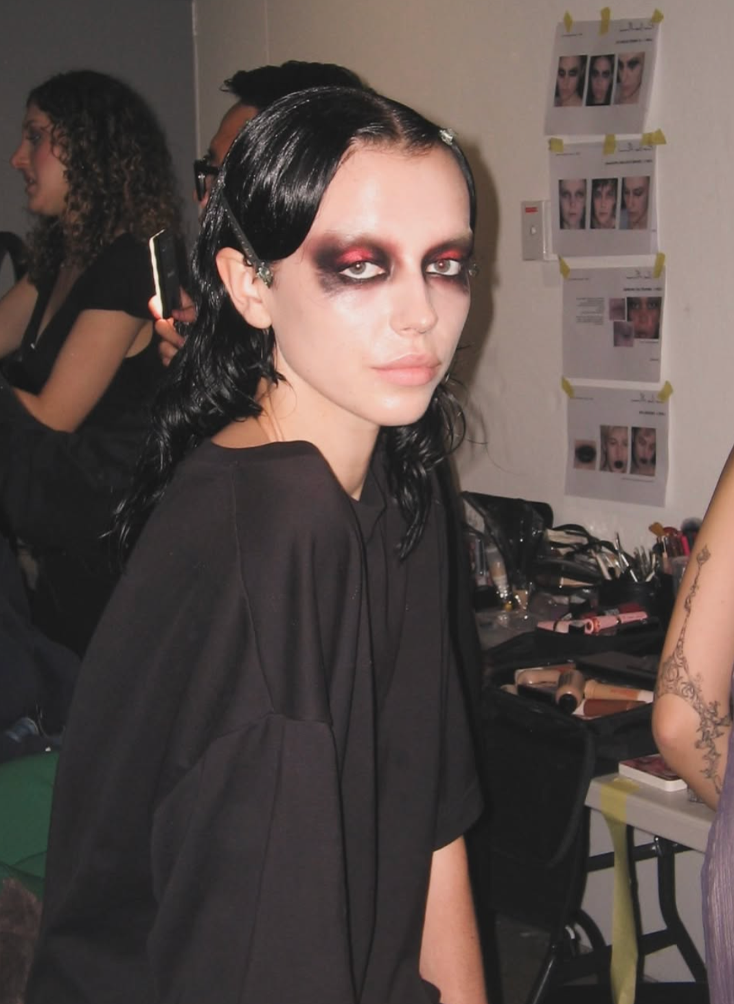 A woman with black curly hair and heavy dark makeup around her eyes, wearing a black top, standing in a backstage or makeup room with makeup products and photos on the wall.