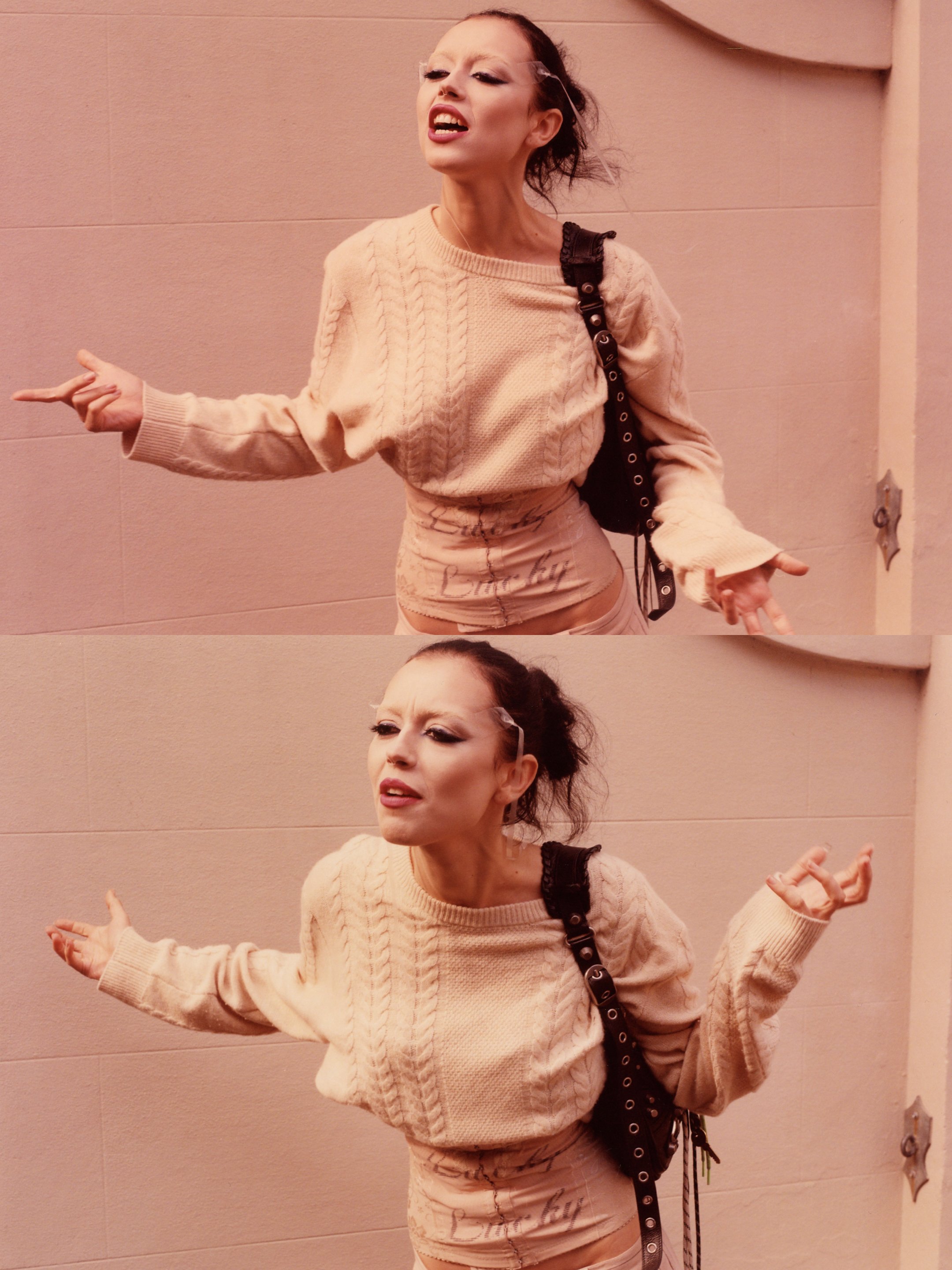 A woman with dark, curly hair styled up and makeup including bold eyeliner and lipstick, standing against a plain wall. She is wearing a beige cable-knit sweater and matching shorts, with a black strap shoulder bag. She appears to be singing or speaking with expressive hand gestures.