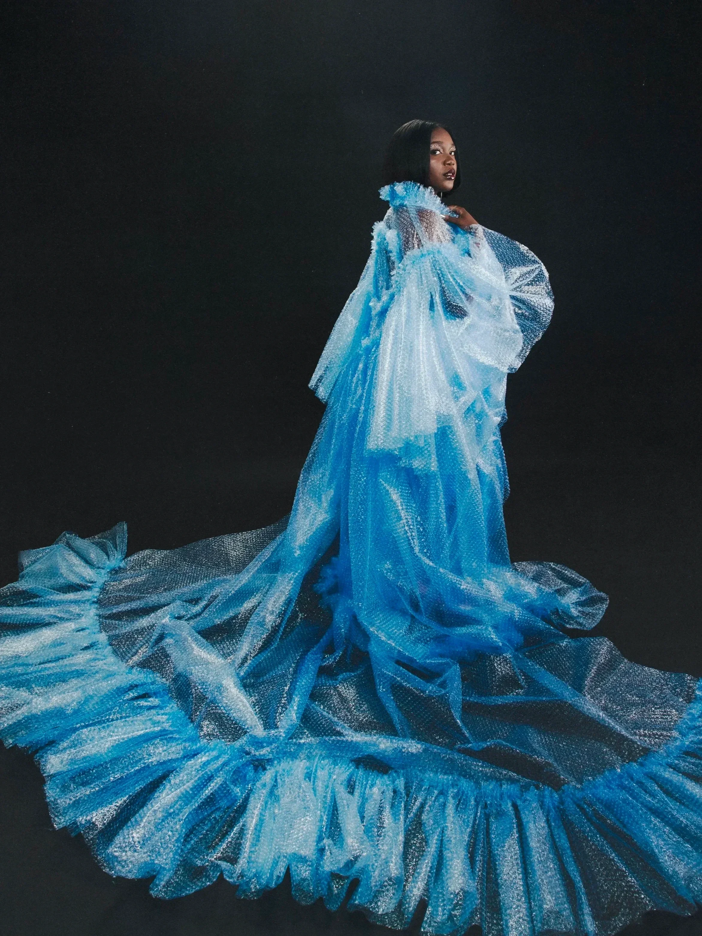A woman with dark hair wearing a flowing, shimmering blue gown against a black background.