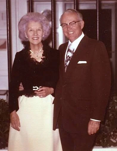 A smiling elderly woman with curly purple hair wearing a black top and white skirt, standing next to a smiling older man with glasses, wearing a dark suit and tie.