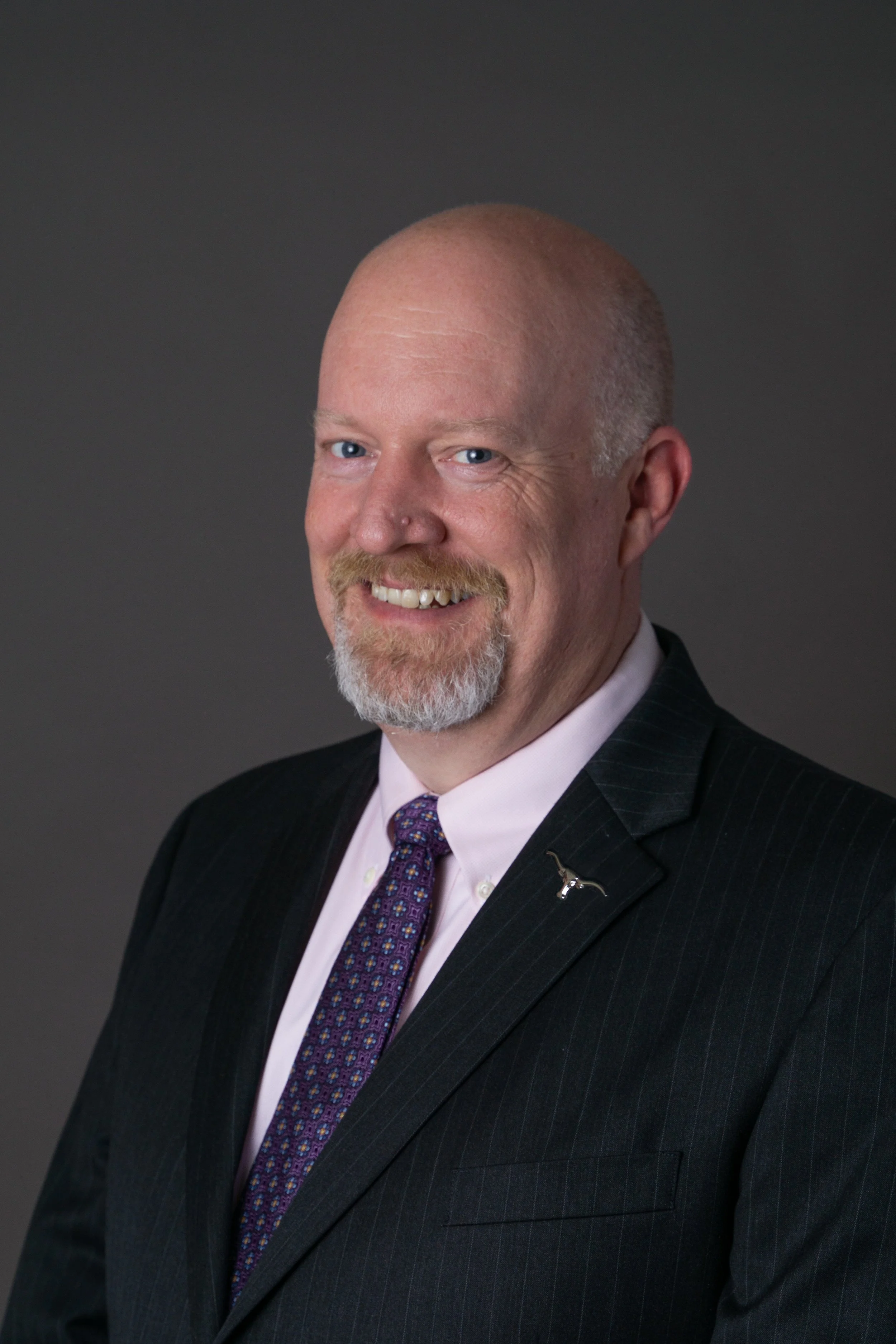 Headshot of a bald man with a beard and mustache, smiling, dressed in a suit and tie.