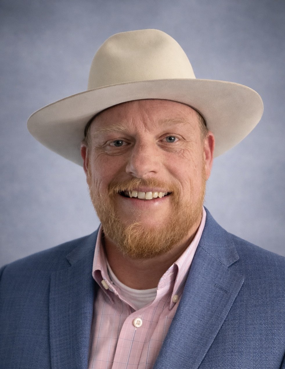 A man with a red beard and mustache wearing a light-colored cowboy hat, a pink dress shirt, and a blue blazer, smiling at the camera with a plain gray background.