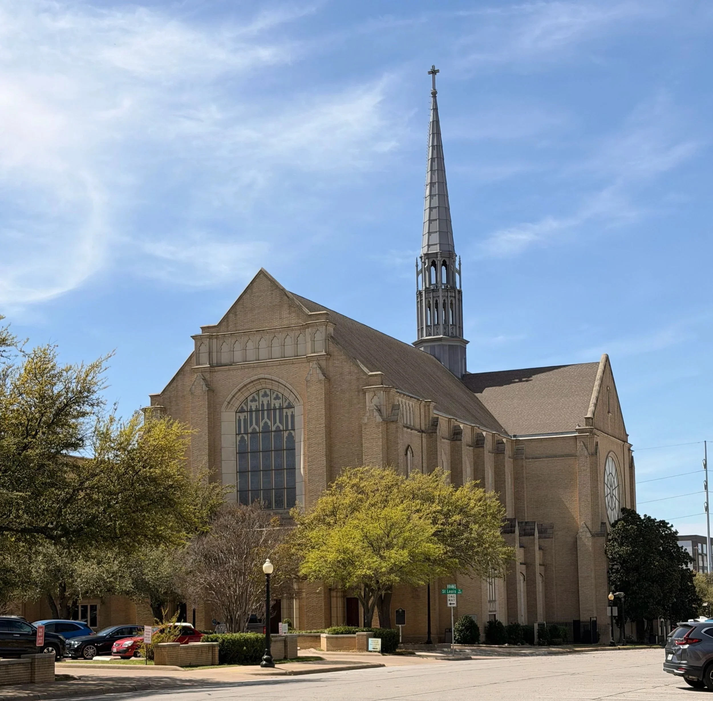 A large church with beige brick walls, tall stained glass windows, and a high steeple with a cross on top, surrounded by trees and parked cars under a partly cloudy sky.