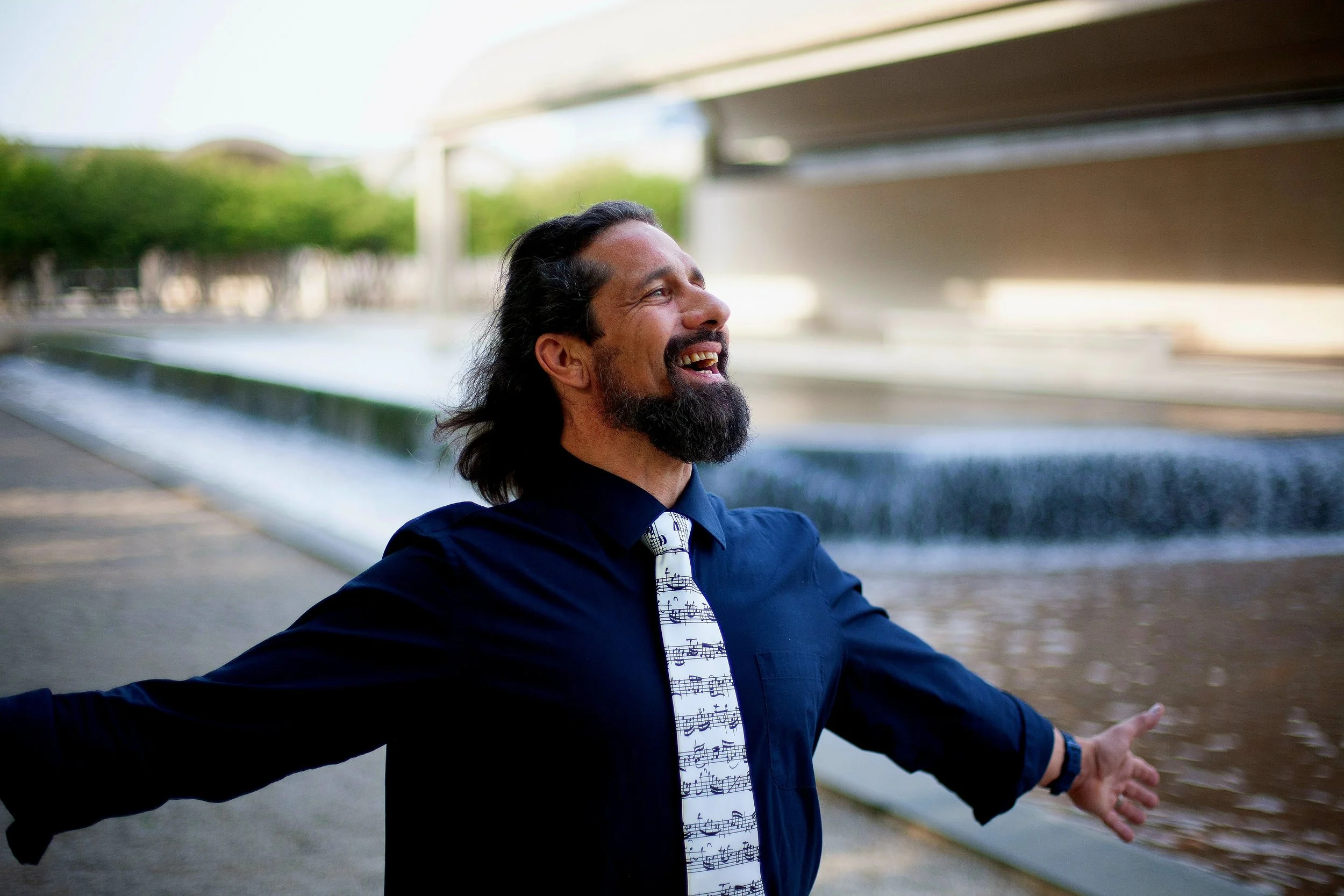 Man with long dark hair and beard smiling with arms outstretched near a modern water feature under a bridge in a park.