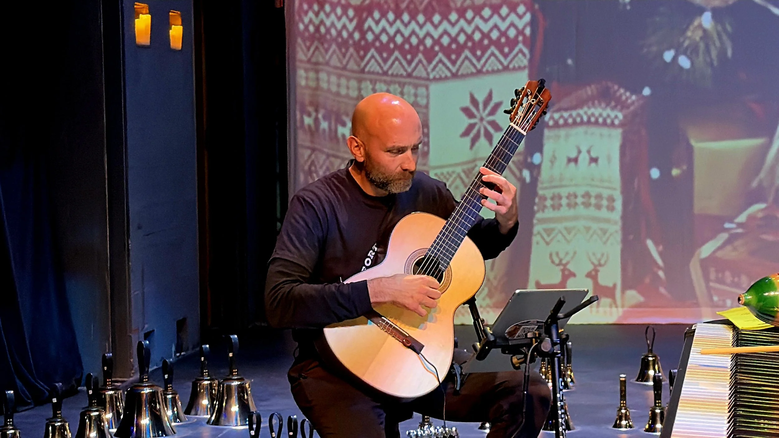 A man with a bald head and beard plays an acoustic guitar on stage, surrounded by various percussion instruments and a keyboard, with a festive holiday backdrop in the background.