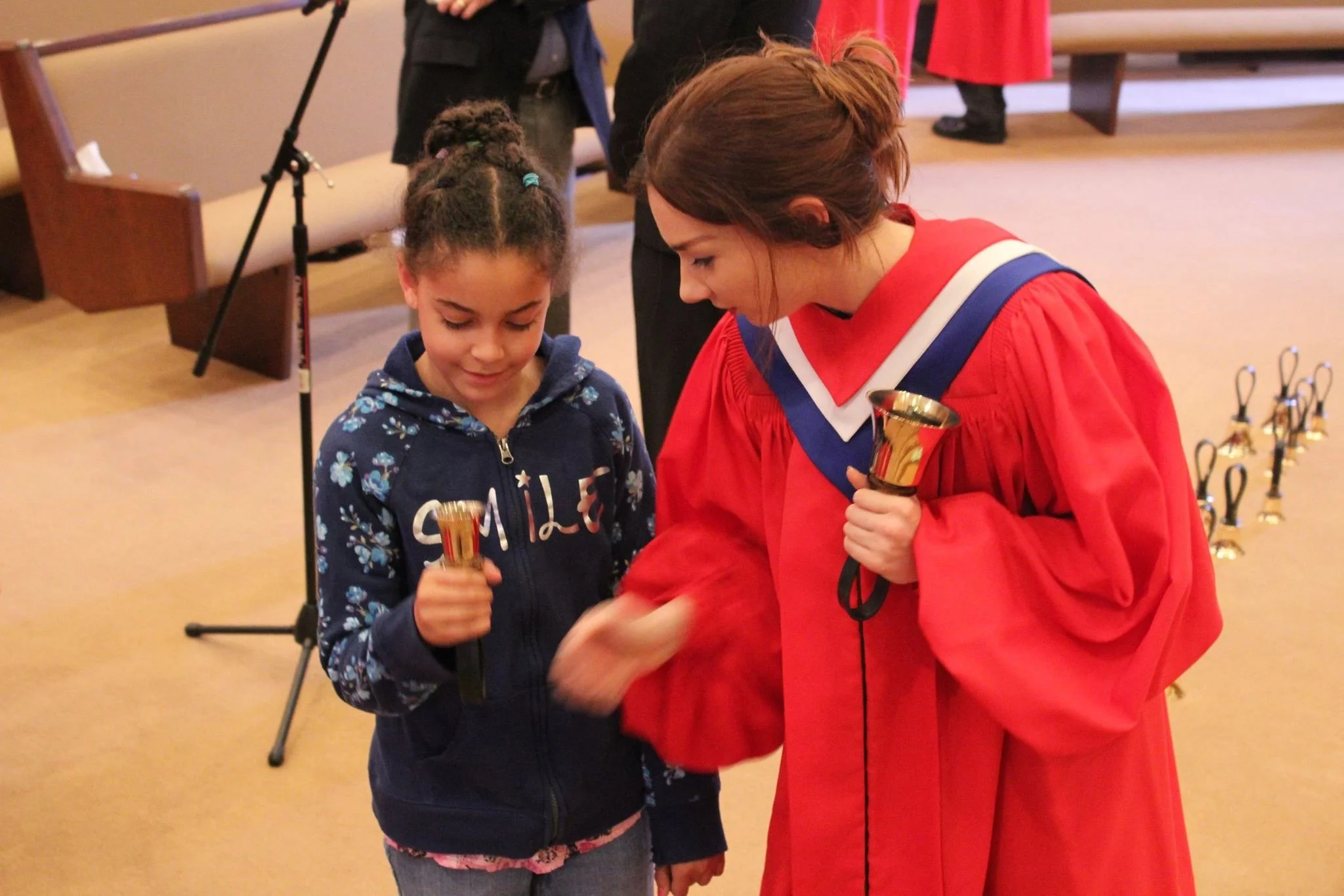 A young girl receiving a medal from a woman in a red academic gown during an award ceremony, both holding small gold trophies, in a room with a beige carpet and benches.