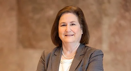 A professional woman with shoulder-length brown hair, wearing a gray blazer and pearl necklace, smiling with arms crossed in front of a blurred brown background.