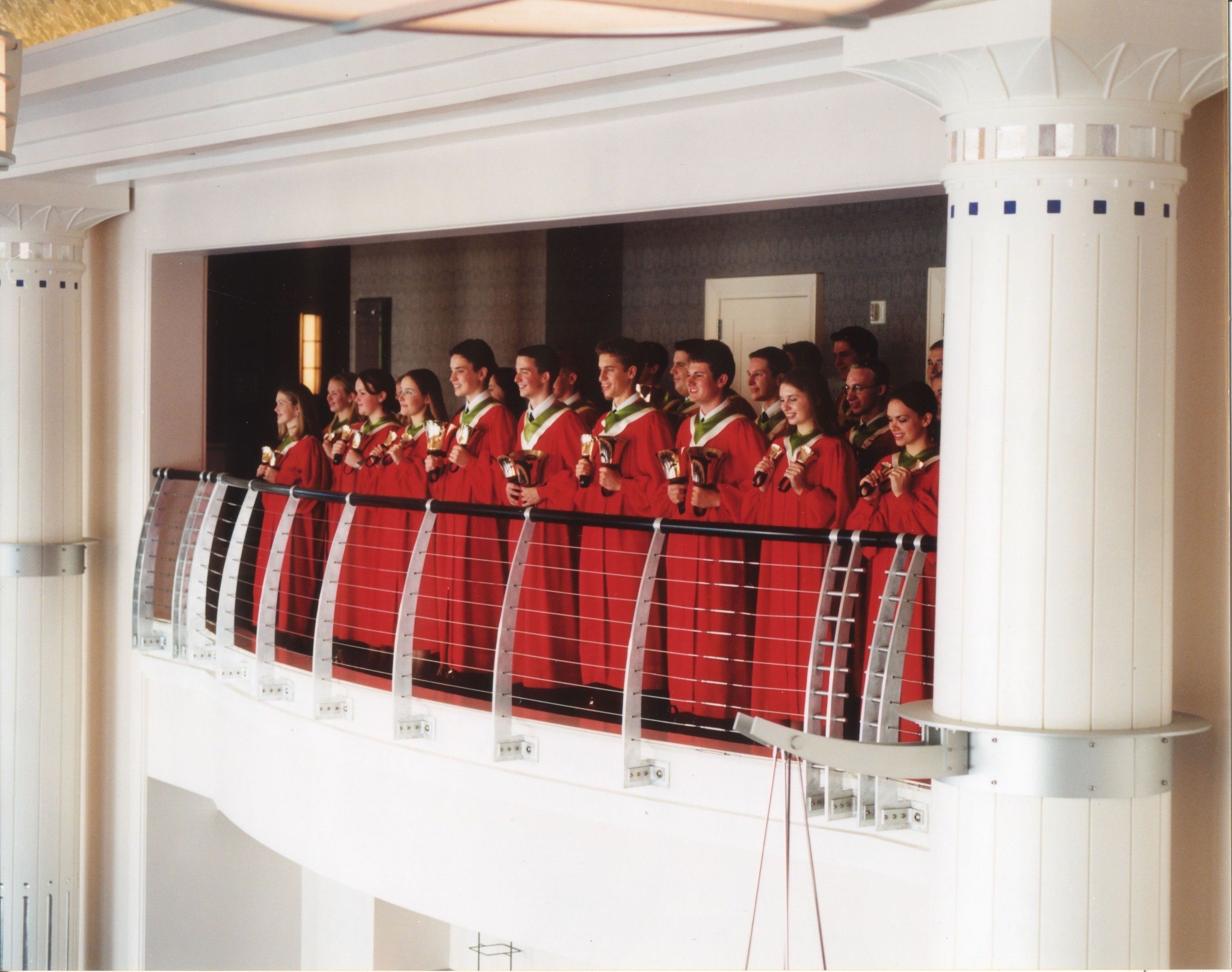 Group of graduates in red gowns holding diplomas, standing on a balcony inside a building.