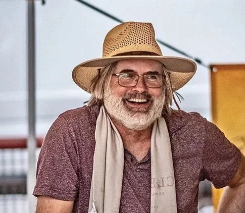 A smiling man with long hair and glasses wearing a straw hat, maroon shirt, and beige scarf outdoors.